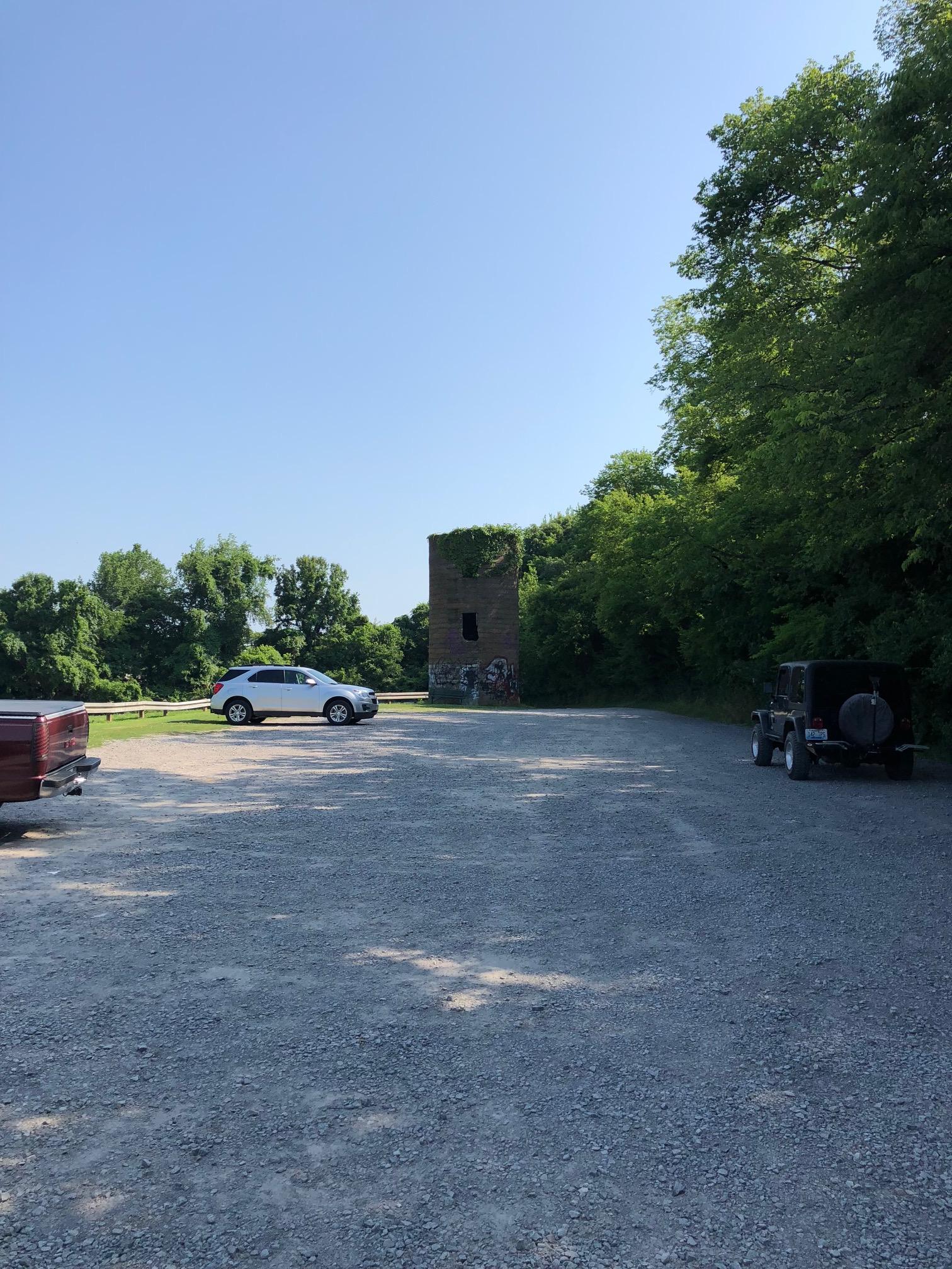 A gravel parking area with two parked vehicles, a red pickup truck and a white SUV, under a clear blue sky. In the background, there is a partially overgrown stone tower with some visible graffiti, surrounded by greenery. Lock 4 mountain bike trail.