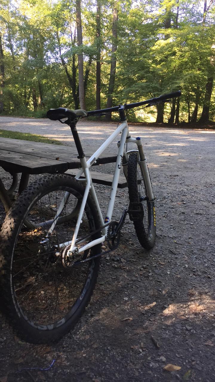 SPOT Rocker SS Steel: A white mountain bike parked beside a wooden picnic table, set against a backdrop of trees and a gravel path. Sunlight filters through the leaves, creating a serene outdoor scene.