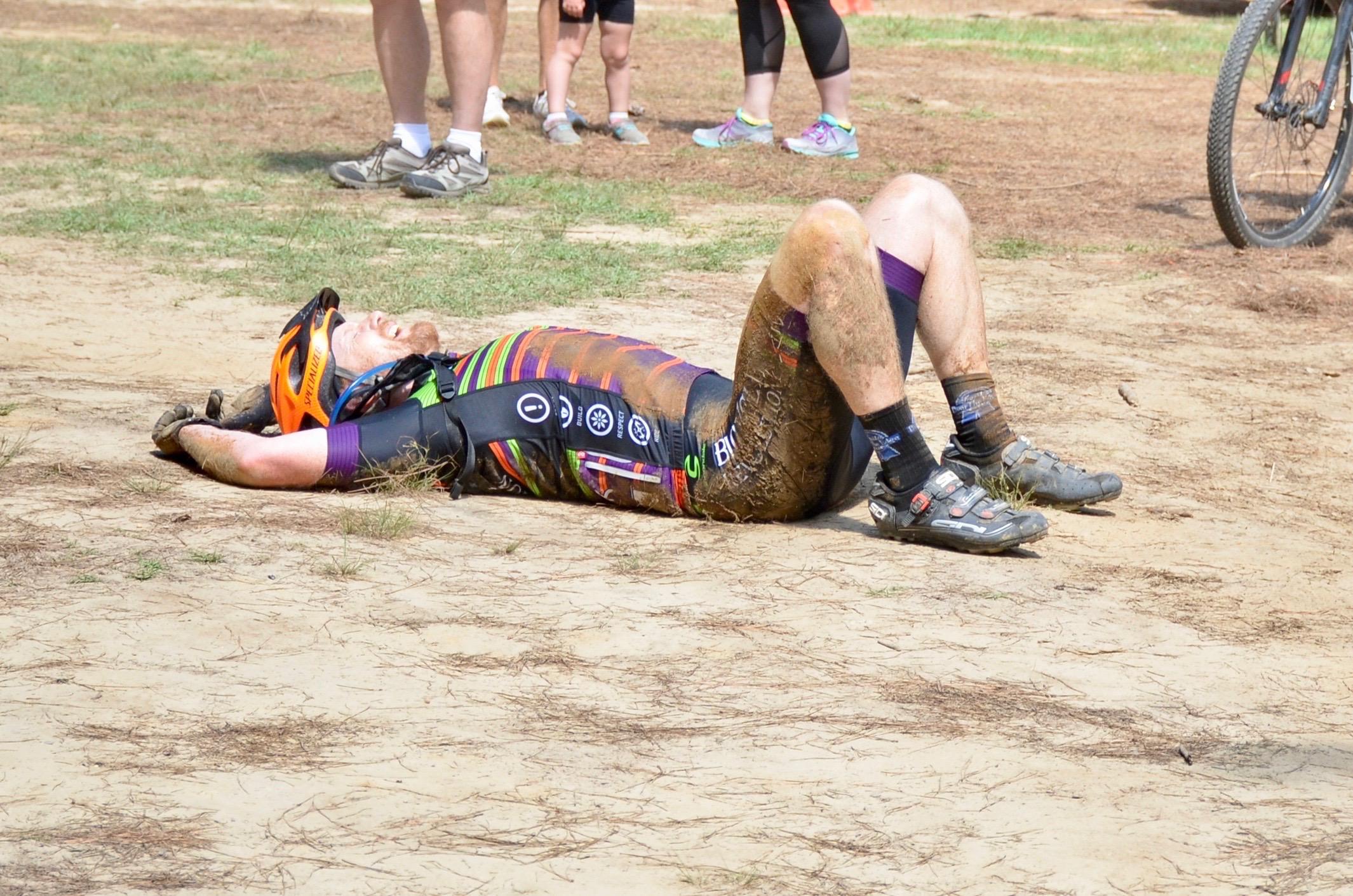 A cyclist lying on the ground after a race, covered in mud and dirt, wearing a colorful cycling outfit and an orange helmet. In the background, people are gathered, some standing and others watching nearby. The scene takes place outdoors, on a dirt surface with patches of grass. Mt. Zion Bike Trails mountain bike trail.