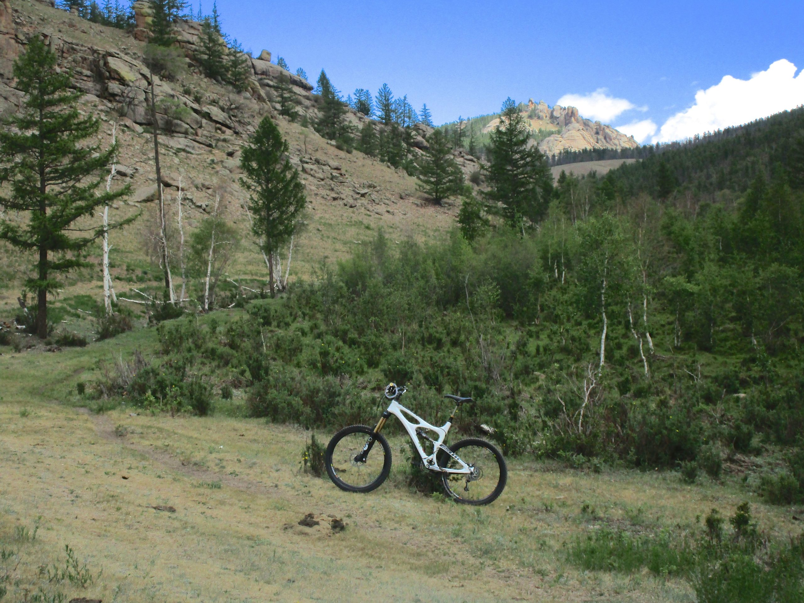 A mountain bike positioned on a grassy trail in a scenic natural landscape, surrounded by green vegetation, rocky hills, and sparse trees under a blue sky with some clouds. Uguumur Trail mountain bike trail.
