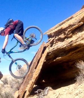 A mountain biker performing a jump off a rocky ledge, with a clear blue sky in the background. The biker is wearing a helmet and cycling gear, showcasing an action-packed moment in a rugged outdoor setting. Wilkins Peak Trails mountain bike trail.