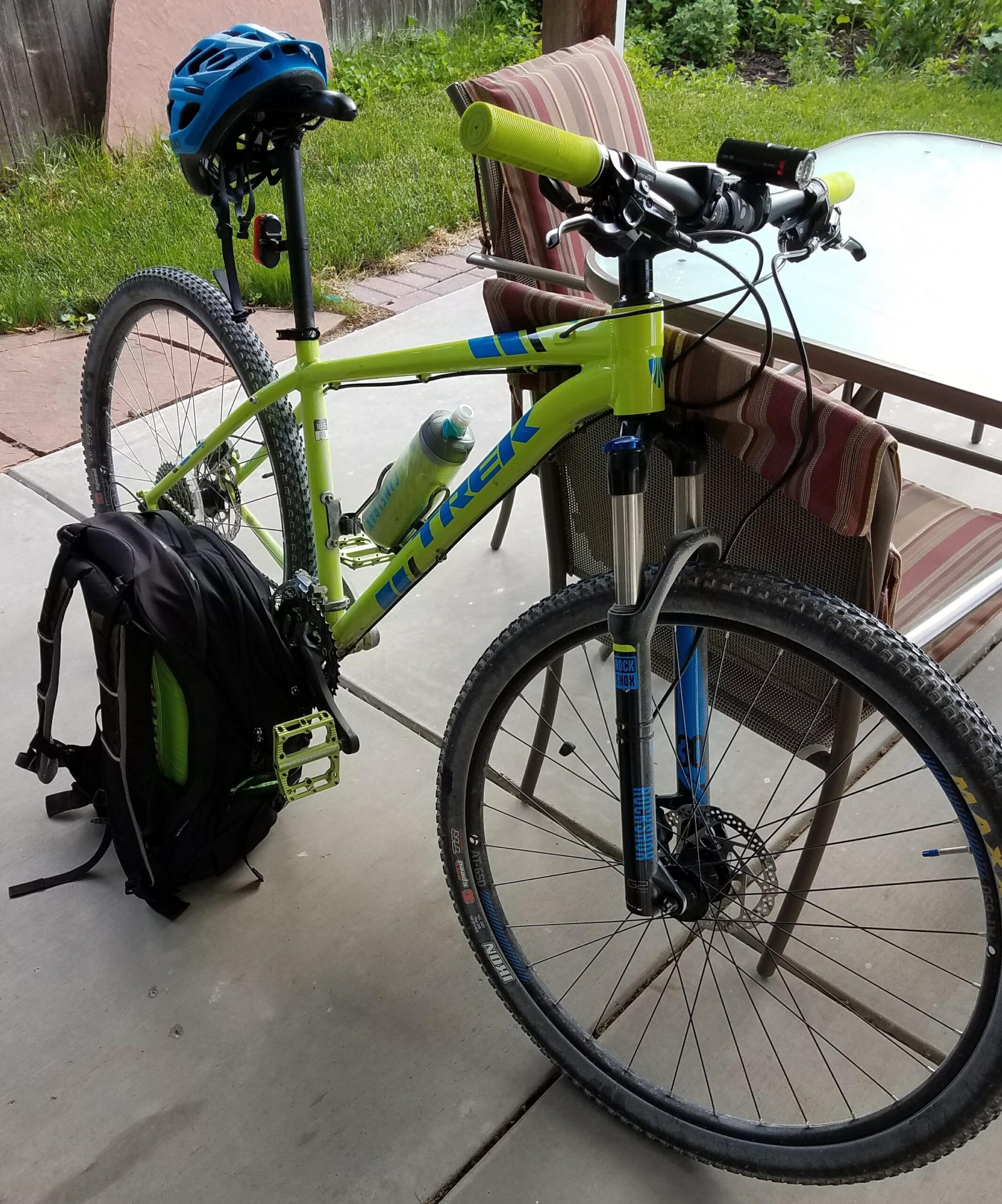 Trek X-Caliber 7: A bright green mountain bike with blue and yellow accents is propped against a black backpack. The bike features a water bottle mounted on the frame, and a blue helmet rests on the seat. In the background, there are grassy areas and a patio table with chairs.