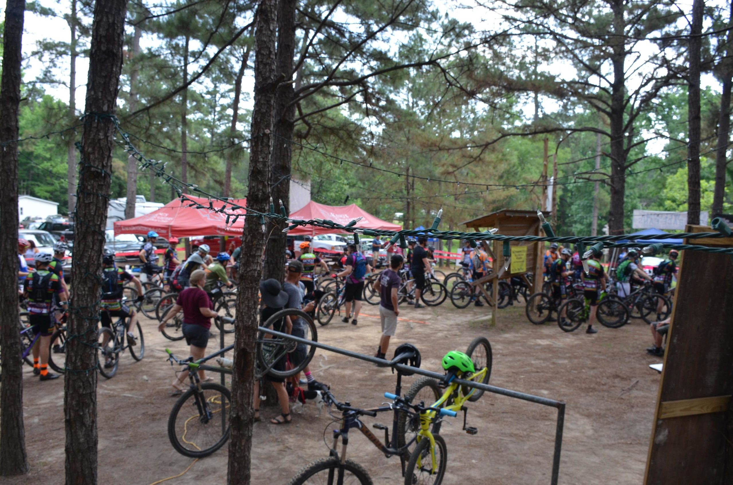 Crowd of mountain bikers gathered in a wooded area, with tents and parked vehicles in the background. Several cyclists are interacting with each other while some bikes are resting on a bike rack. The scene is vibrant, showcasing the outdoor atmosphere of a biking event. Mt. Zion Bike Trails mountain bike trail.
