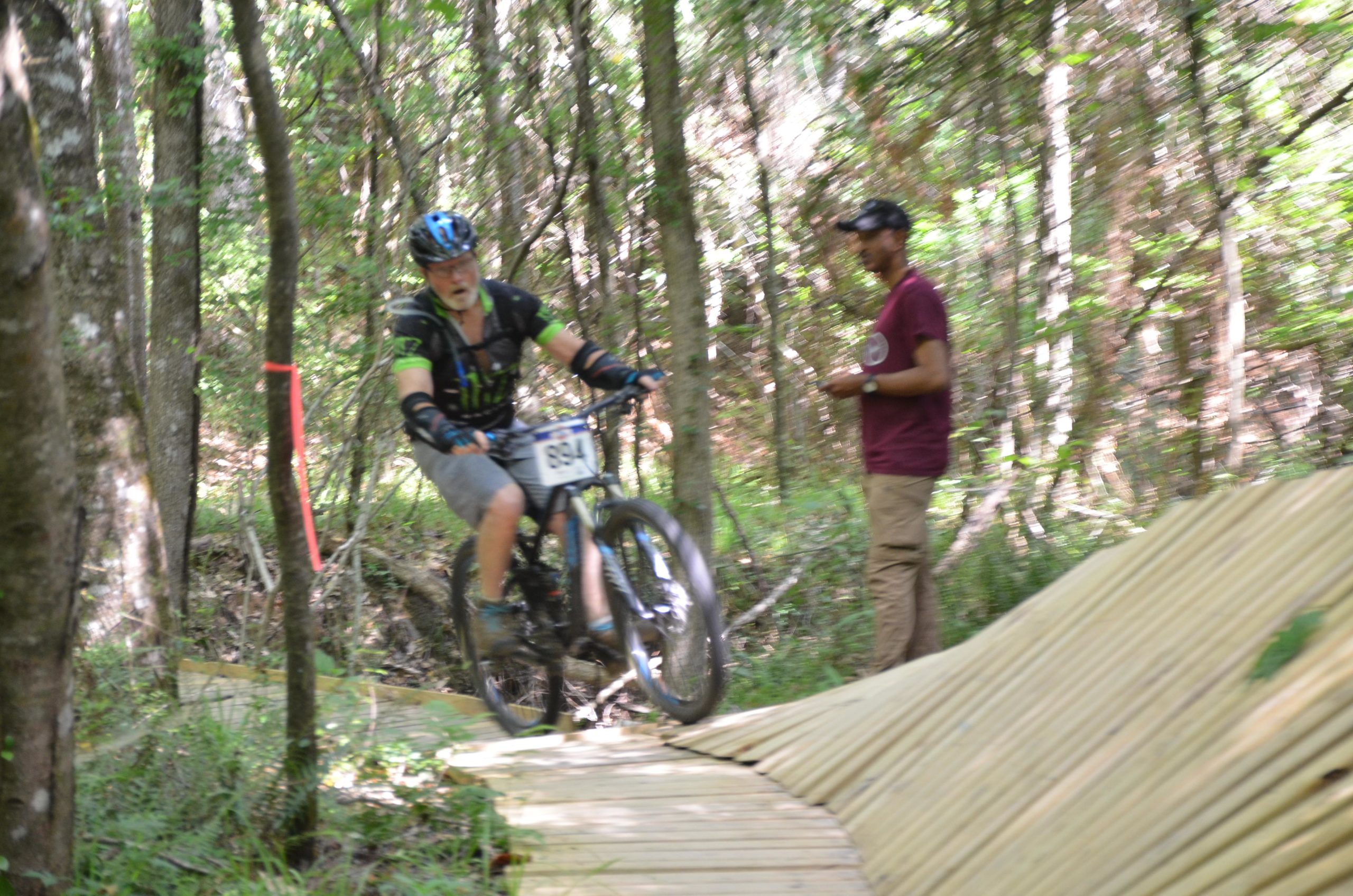 A mountain biker with a number plate rides on a wooden trail through a wooded area, while a bystander watches nearby. Mt. Zion Bike Trails mountain bike trail.
