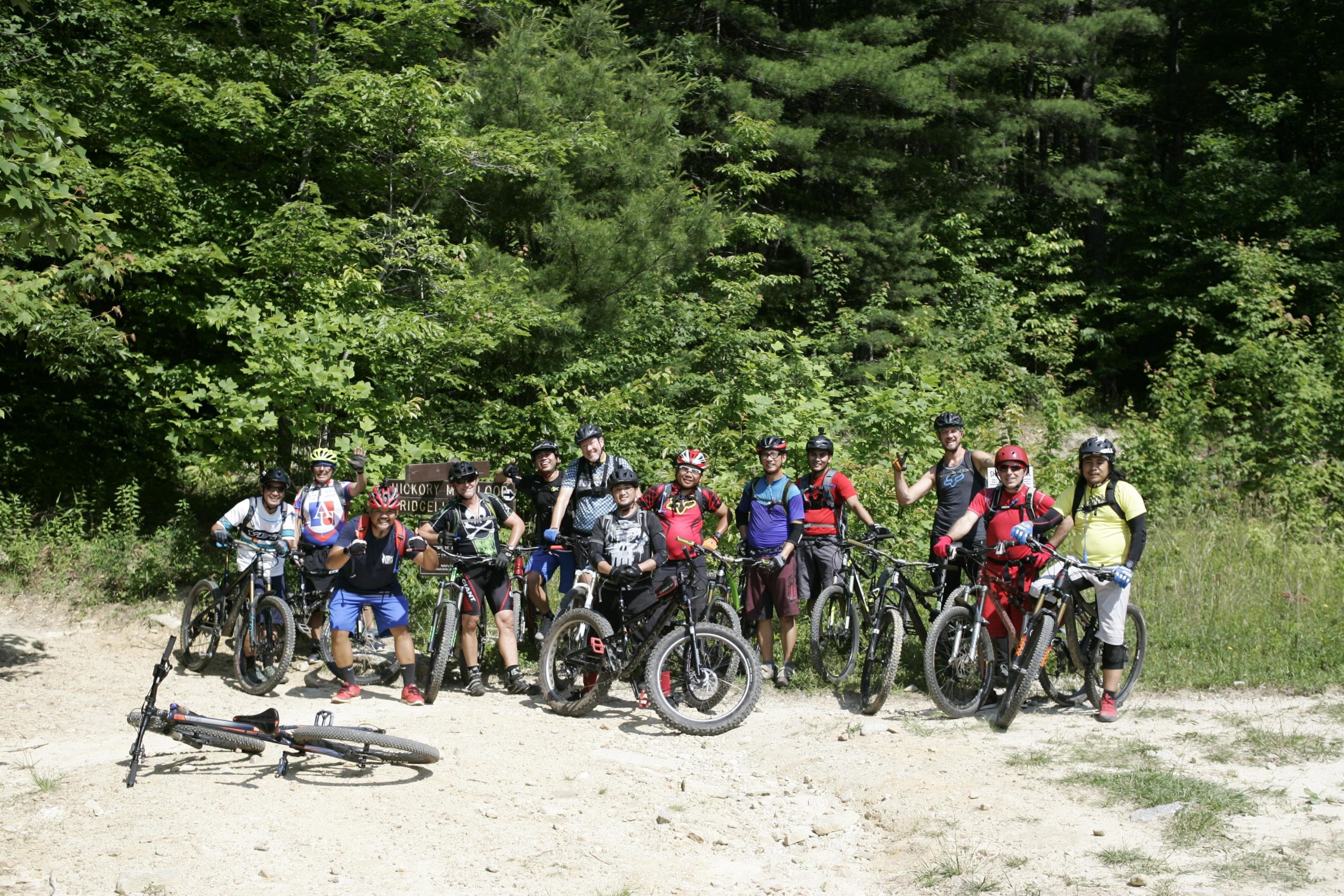 A diverse group of mountain bikers posing together with their bikes in a forested area. The group is smiling and gesturing playfully, showcasing a sense of camaraderie. In the foreground, a bike lies on the ground, while lush green trees surround the scene. DuPont State Forest mountain bike trail.