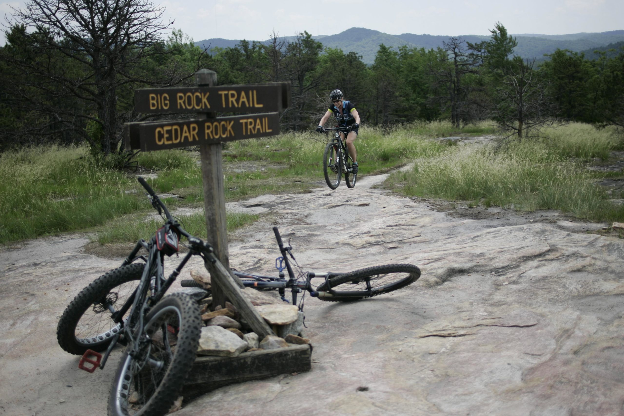 A mountain biker performs a jump on a rocky trail near a signpost pointing to "Big Rock Trail" and "Cedar Rock Trail." Two mountain bikes are leaned against the sign, with a landscape of trees and rolling hills in the background. Big Rock Trail #3 mountain bike trail.