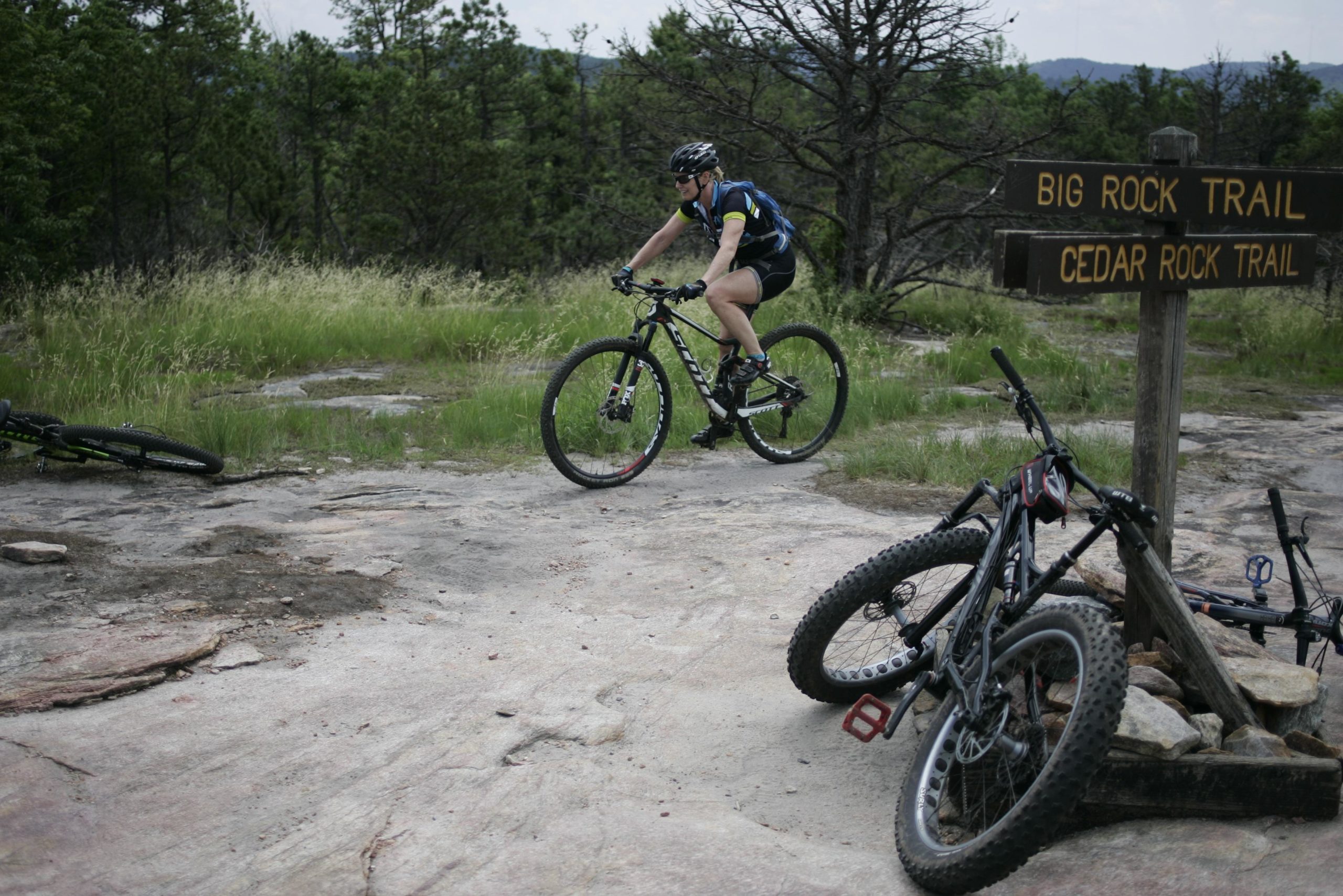 A mountain biker riding on a rocky trail, with a signpost indicating "Big Rock Trail" and "Cedar Rock Trail" nearby. Two bicycles are leaned against a wooden structure on the ground. The surrounding area features grassy patches and sparse trees. DuPont State Forest mountain bike trail.