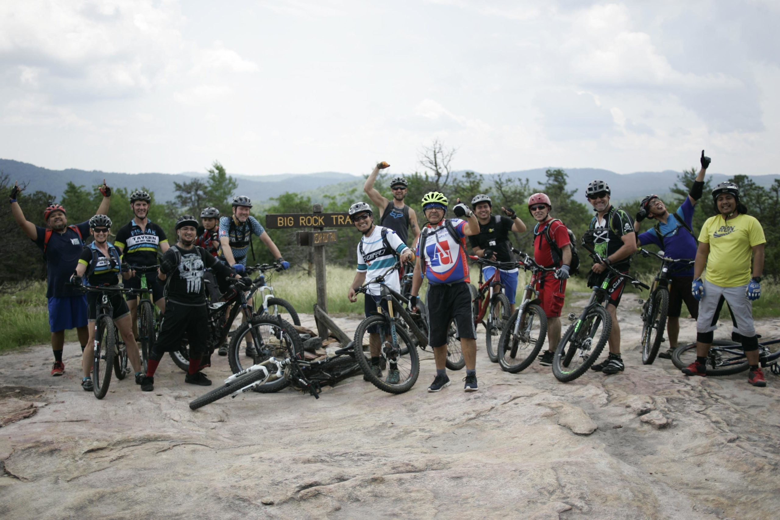 A group of mountain bikers poses joyfully on a rocky outcrop near a trail sign that reads "Big Rock Trail." The riders, wearing helmets and biking gear, display enthusiastic gestures, with bicycles parked beside them. Lush green trees and rolling hills are visible in the background under a partly cloudy sky. Big Rock Trail #3 mountain bike trail.