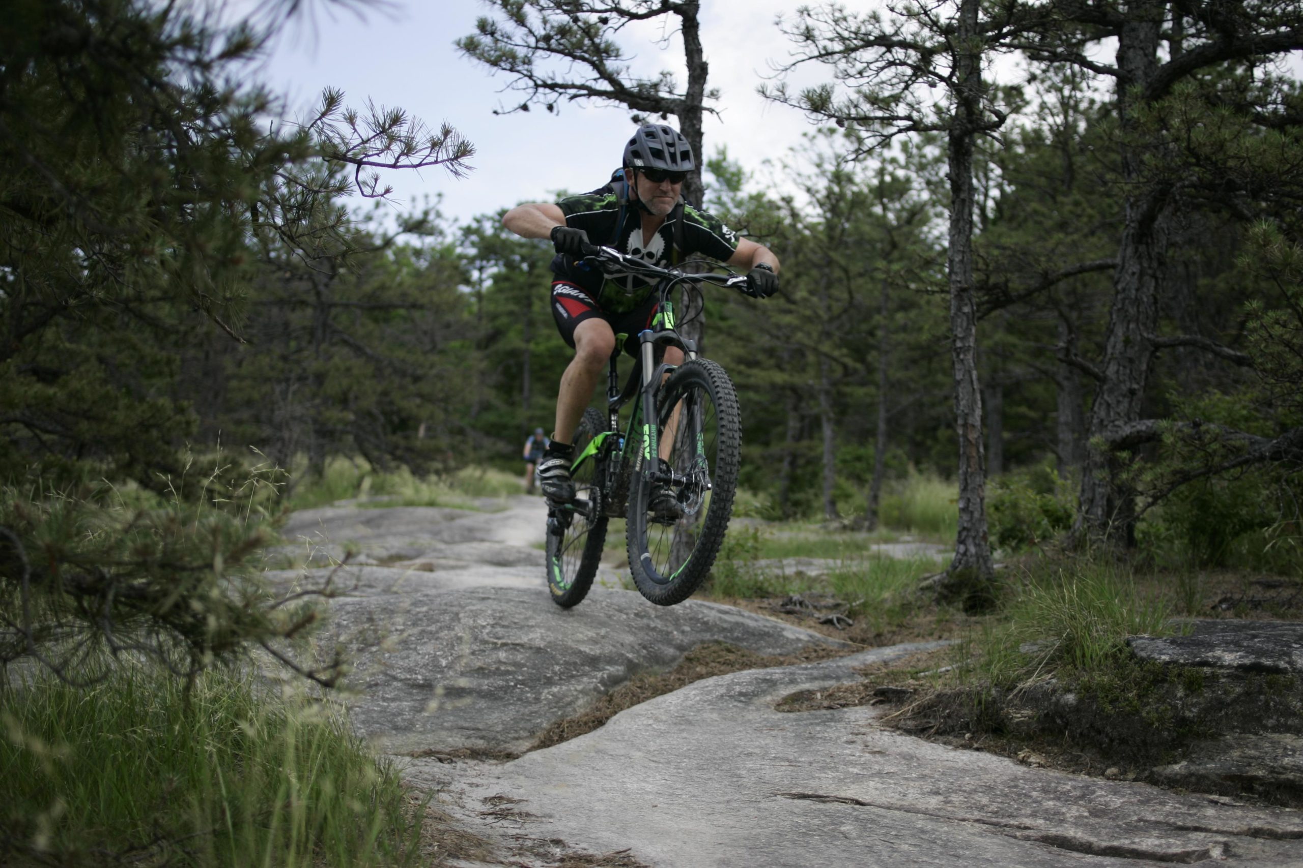 A person in a black helmet and sports gear riding a green mountain bike, performing a jump over a rocky trail surrounded by trees and greenery. Big Rock Trail #3 mountain bike trail.