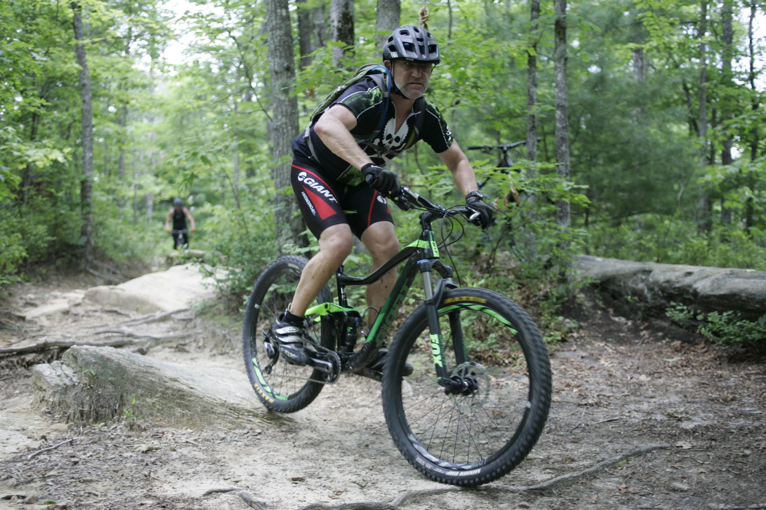 A cyclist riding a mountain bike down a rocky trail in a wooded area, with lush green trees surrounding the path. The rider is wearing a helmet and biking gear, demonstrating an active and adventurous lifestyle. In the background, another cyclist can be seen navigating the trail. Big Rock Trail #3 mountain bike trail.