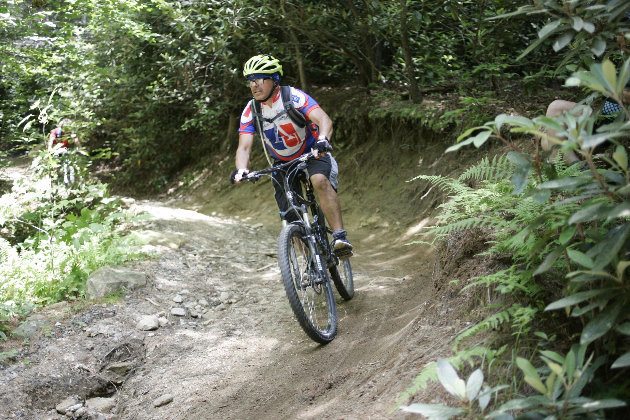 A cyclist riding a mountain bike on a dirt trail surrounded by lush greenery and ferns. The rider is wearing a bright helmet and a colorful shirt, focused on navigating the trail. Big Rock Trail #3 mountain bike trail.