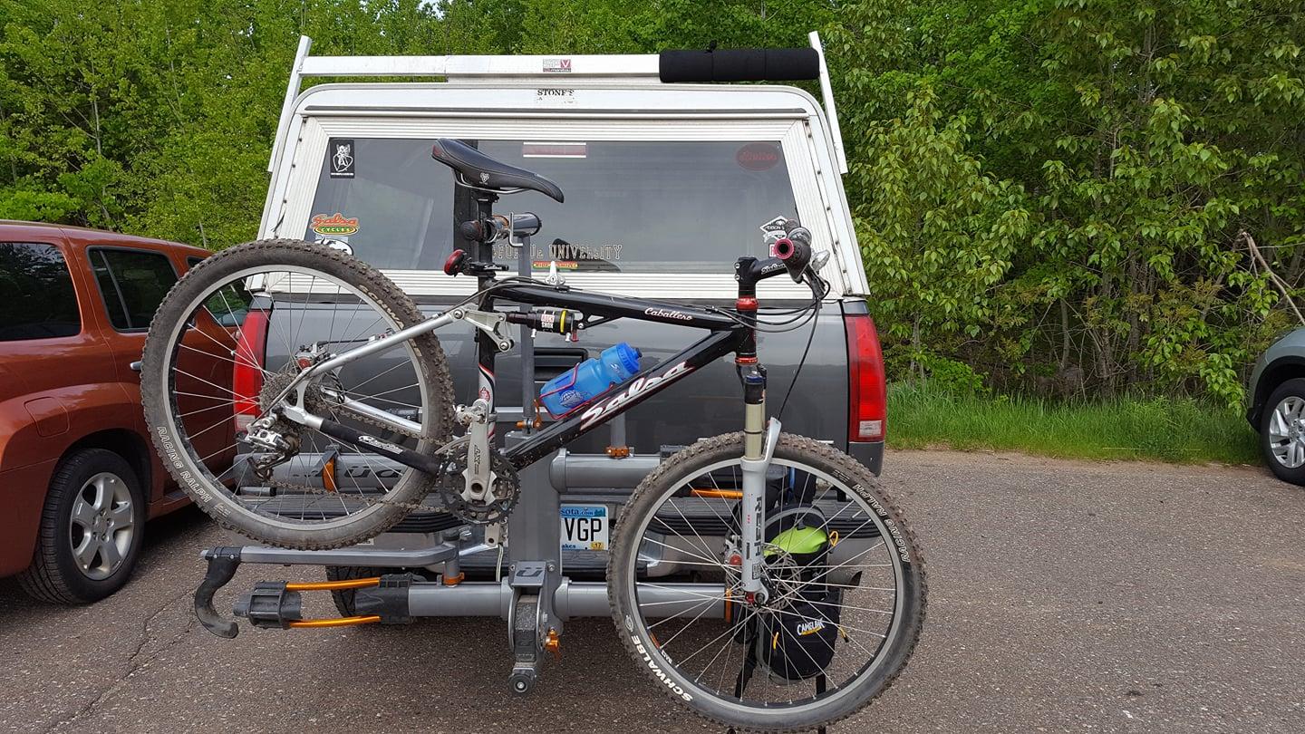 Salsa Caballero: A mountain bike is mounted on a rack attached to the back of a vehicle, parked in a natural setting with greenery in the background. A water bottle is secured in the bike