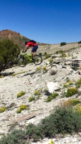 A mountain biker in a red shirt and blue shorts is mid-jump over rocky terrain, with a clear blue sky and scattered vegetation in the background. Wilkins Peak Trails mountain bike trail.