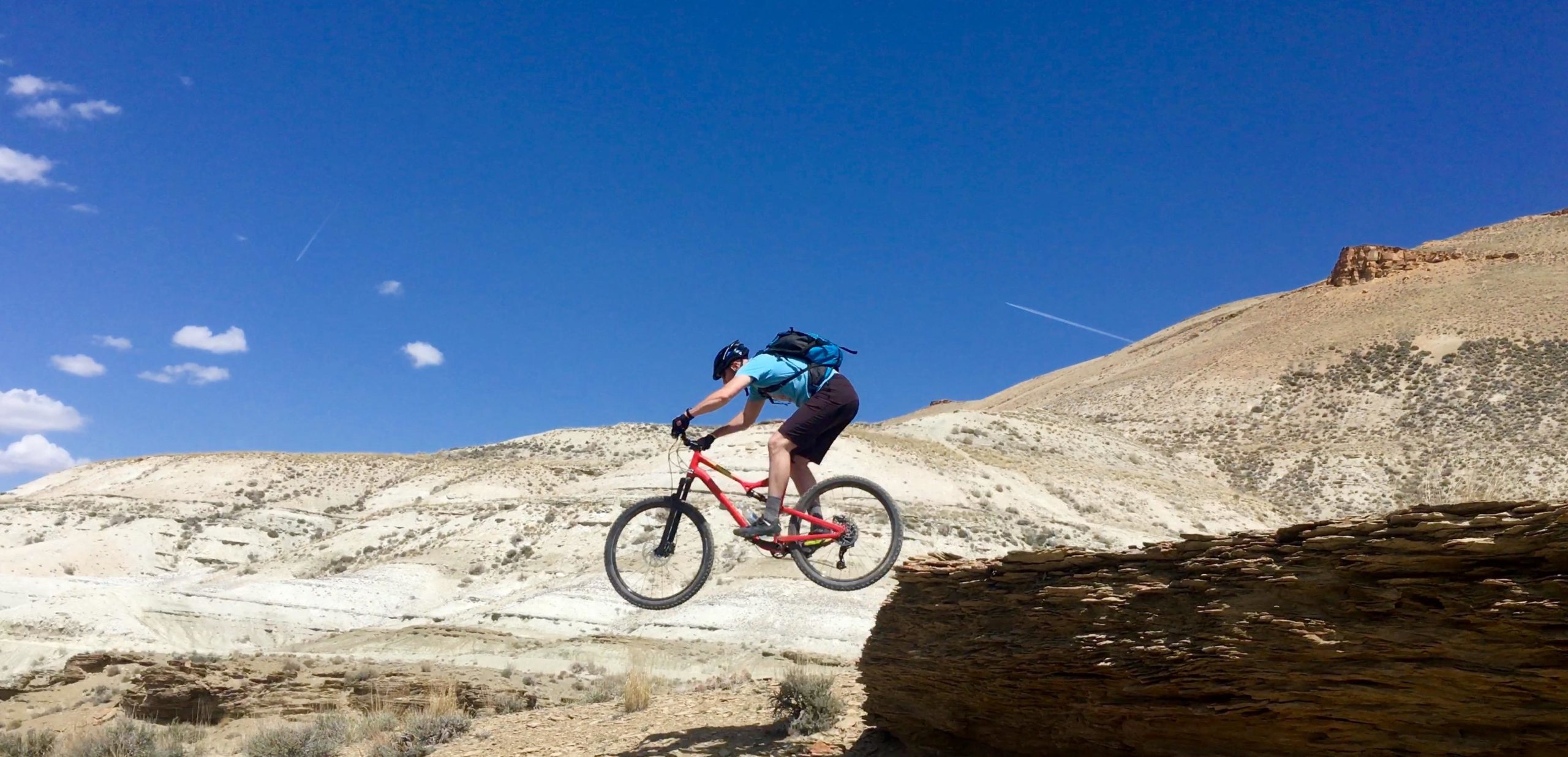 A mountain biker in a blue shirt and helmet jumps off a rock ledge, soaring through the air over a picturesque, rugged landscape under a clear blue sky with a few clouds. The terrain features dry hills and sparse vegetation, highlighting an adventurous outdoor setting. Wilkins Peak Trails mountain bike trail.