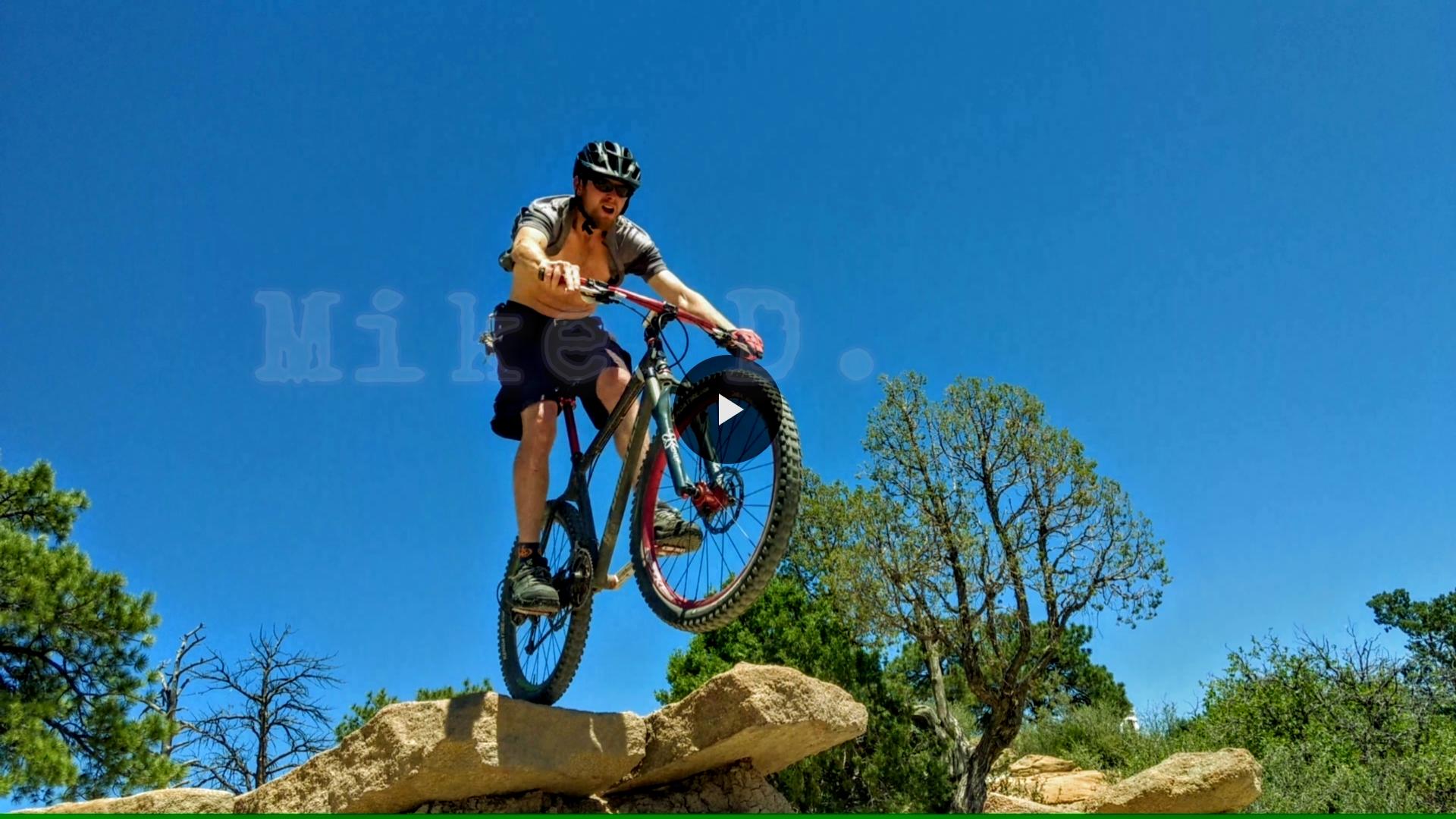 Alt text: A shirtless man in a bike helmet performs a jump on a mountain bike, with his front wheel elevated above a rocky ledge. The background features a clear blue sky and green trees. Palmer Park mountain bike trail.