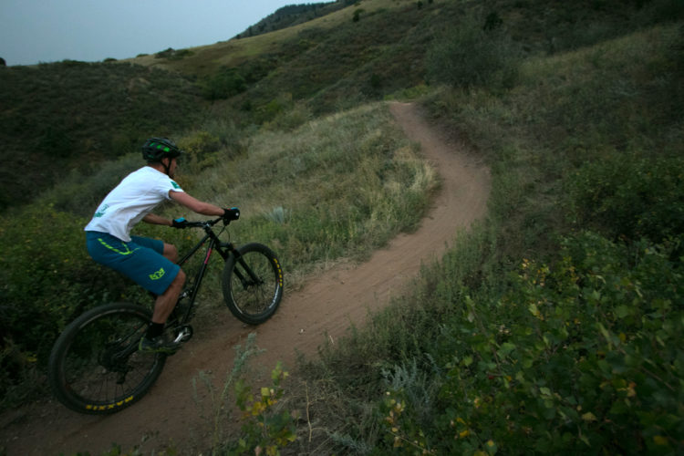 A mountain biker navigating a curving dirt trail through a green hillside, wearing a helmet and cycling gear, with an emphasis on the natural landscape and vegetation surrounding the path.