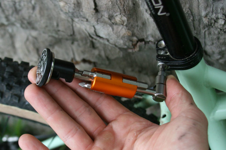 A hand holding a multi-tool with an orange center section and various screwdriver tips, placed next to a bicycle's handlebar. The background features a textured rock surface and a bicycle tire, indicating an outdoor setting.