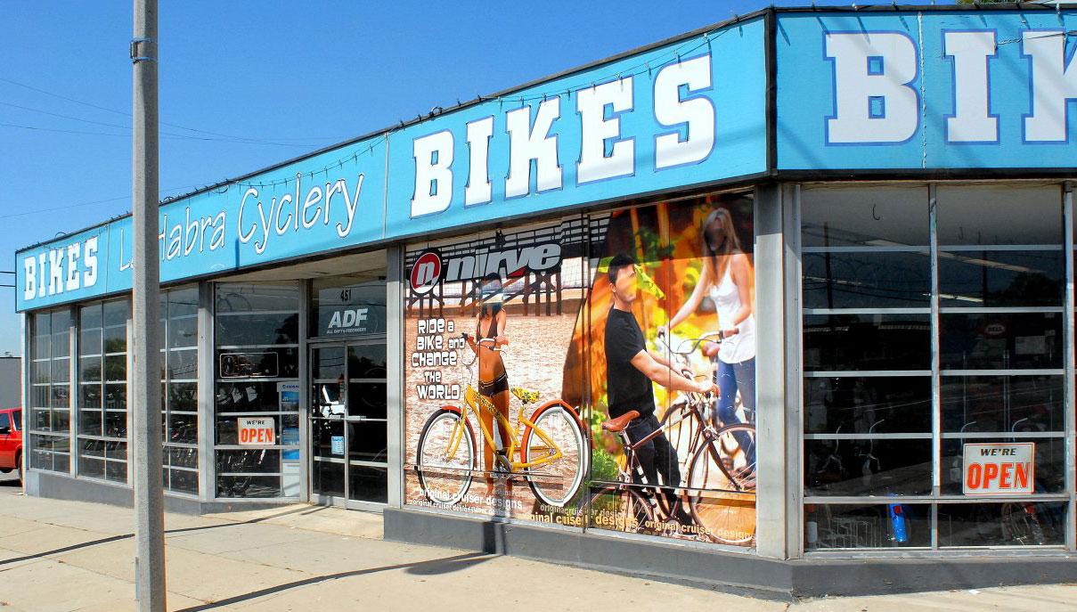 Exterior view of La Habra Cyclery, a bike shop, featuring a bright blue sign that reads "Bikes." The storefront displays an advertisement with two people on bicycles, promoting a biking lifestyle. Clear glass windows showcase various bicycles inside, and an open sign is visible, indicating the shop is open for business.