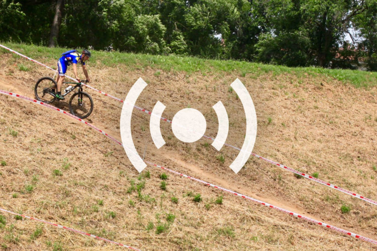 A mountain biker navigates a slope lined with tape during a race, surrounded by grassy terrain and trees in the background.