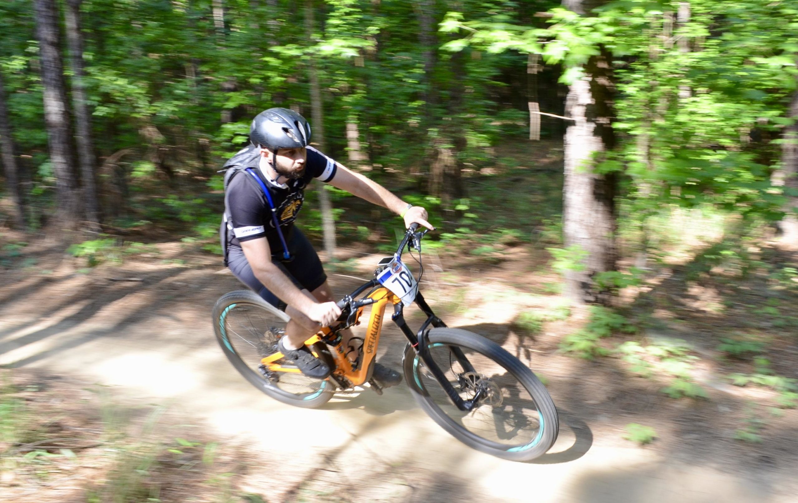 A person wearing a helmet and cycling gear rides a mountain bike quickly along a dirt trail in a wooded area, surrounded by greenery. The bike is an orange and black model, and the rider appears focused and in motion. Mt. Zion Bike Trails mountain bike trail.