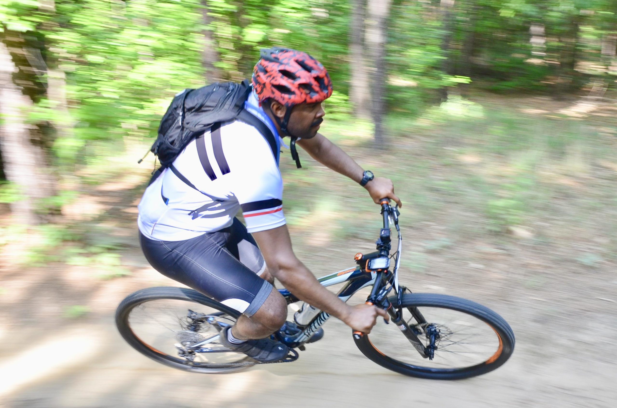 A person riding a mountain bike on a dirt trail surrounded by lush green trees, wearing a helmet and athletic clothing, with a backpack. The image captures the sense of speed and motion, with a blurred background emphasizing the outdoor adventure. Mt. Zion Bike Trails mountain bike trail.