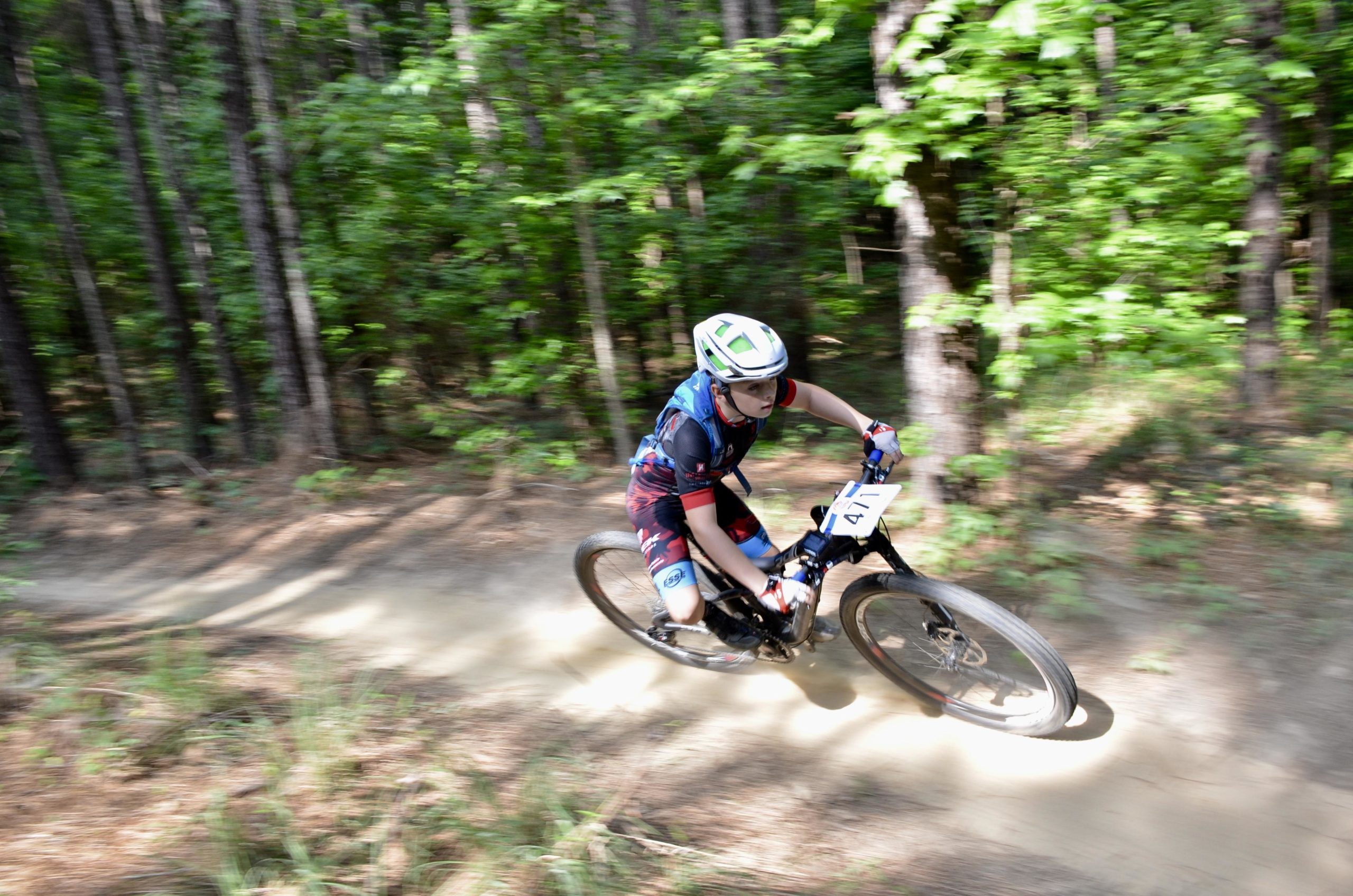 A young cyclist races along a dirt trail in a forest, kicking up dust as they navigate a sharp turn. The rider, wearing a helmet and colorful cycling gear, is focused on maintaining speed and balance amid the trees. Mt. Zion Bike Trails mountain bike trail.