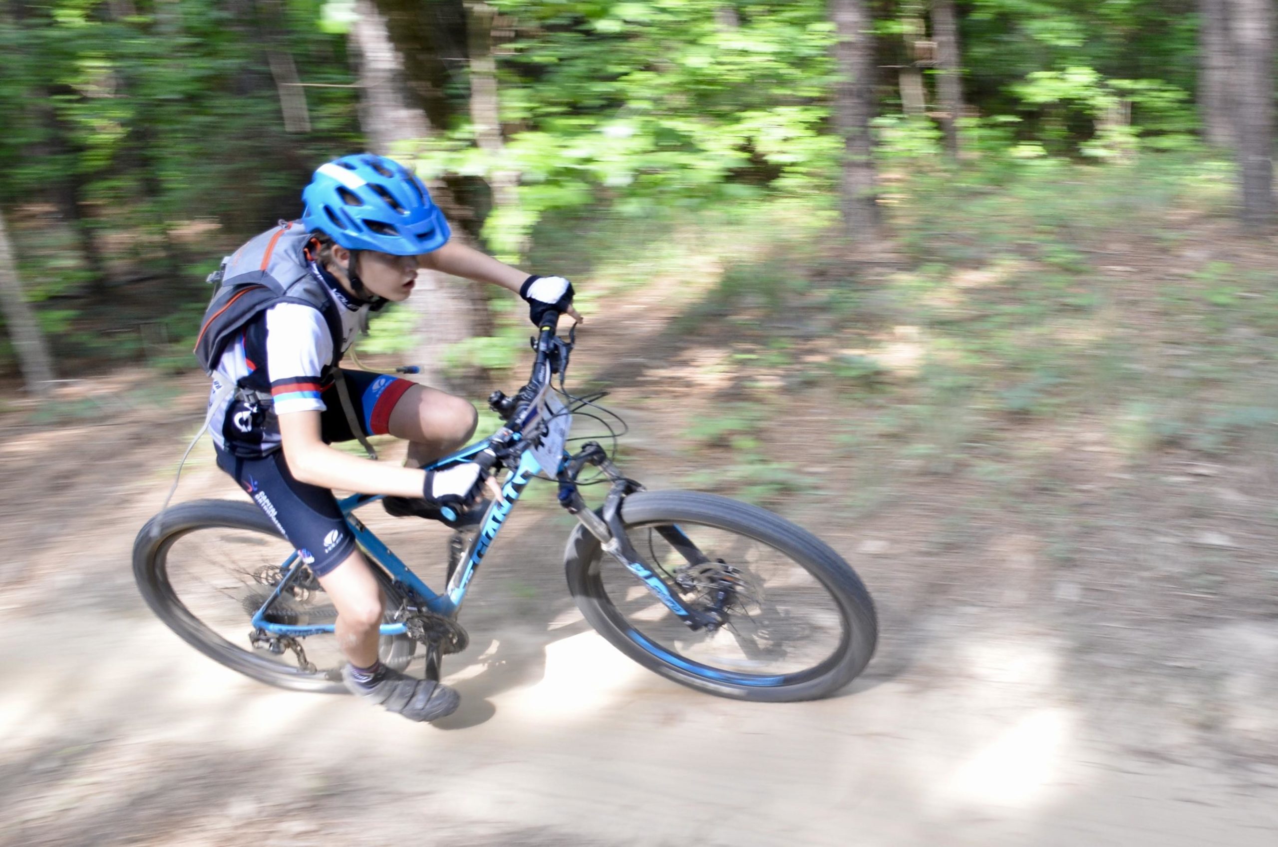 A young cyclist wearing a blue helmet and sports attire is navigating a dirt trail on a mountain bike, captured in motion with a slight blur to emphasize speed. The surrounding greenery and trees create a natural, outdoor setting. Mt. Zion Bike Trails mountain bike trail.