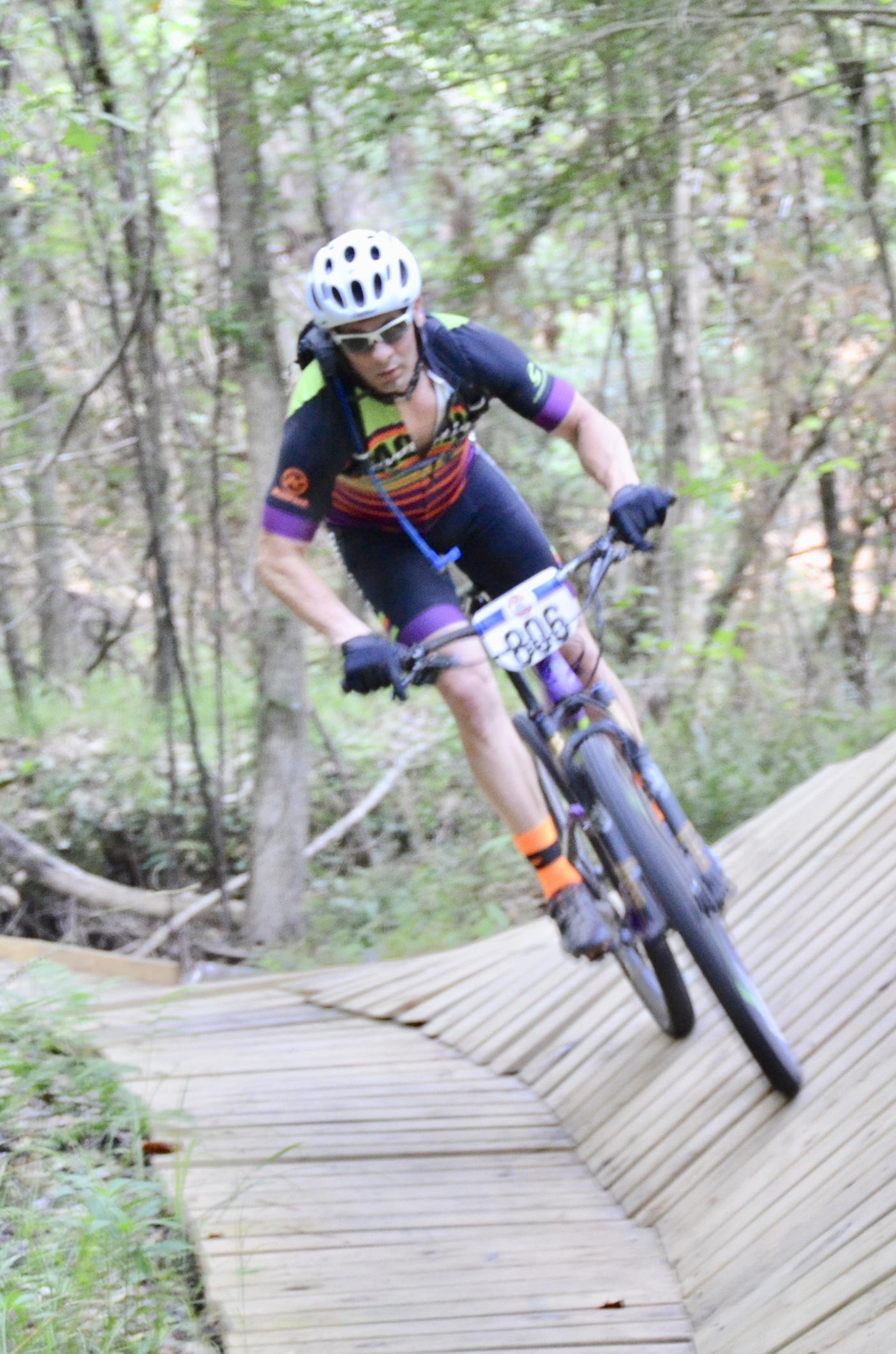 A mountain biker wearing a helmet and colorful cycling gear is navigating a wooden trail in a forested area. The cyclist is leaning into a turn while riding a mountain bike on a sloped wooden surface, surrounded by trees and greenery. Mt. Zion Bike Trails mountain bike trail.
