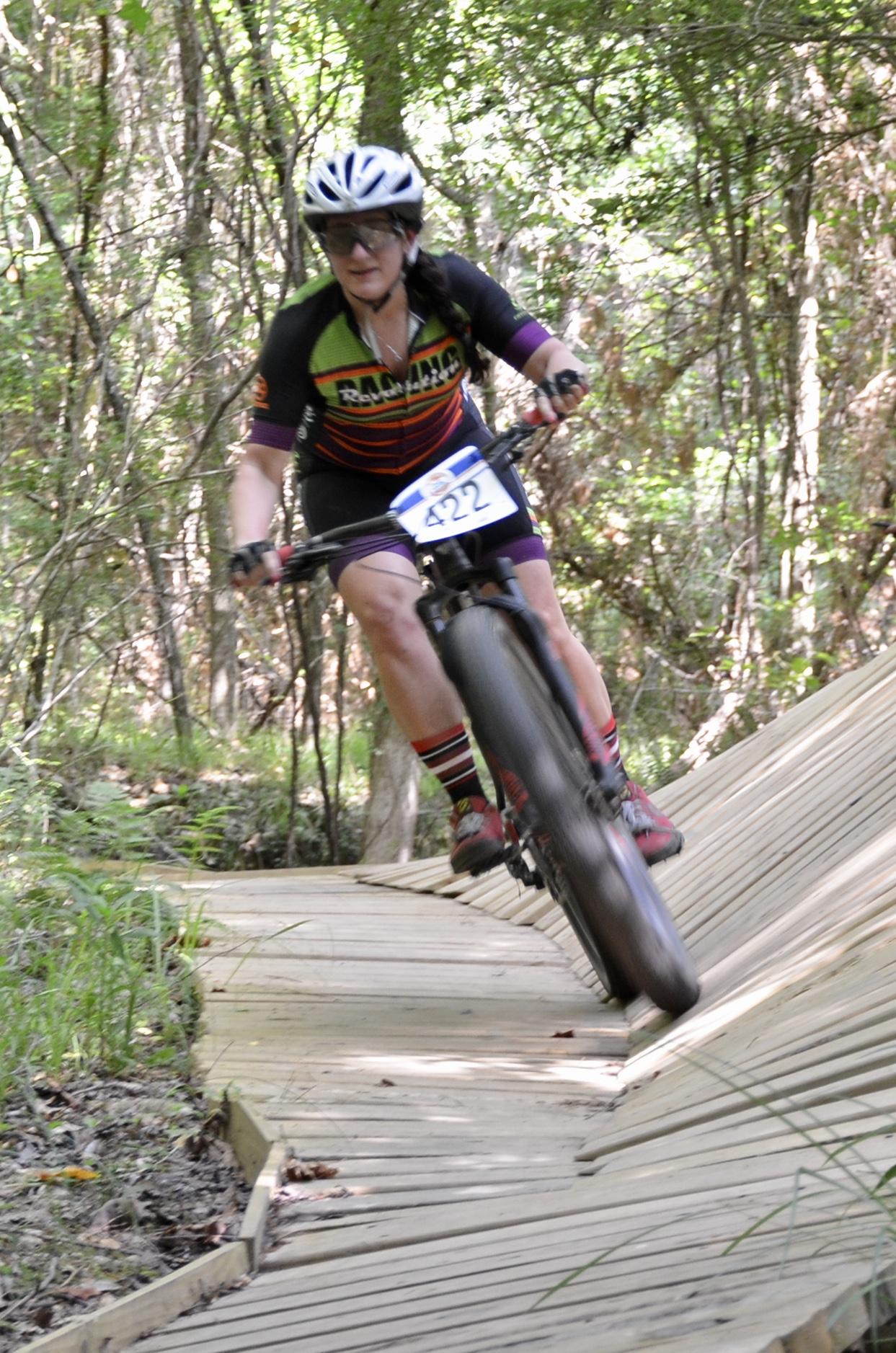 A cyclist in a helmet and sunglasses navigates a wooden biking trail through a forest, showcasing focused determination. The cyclist wears a colorful jersey with the number 422 displayed prominently. The scene captures the dynamic movement and nature of mountain biking, surrounded by greenery and trees. Mt. Zion Bike Trails mountain bike trail.