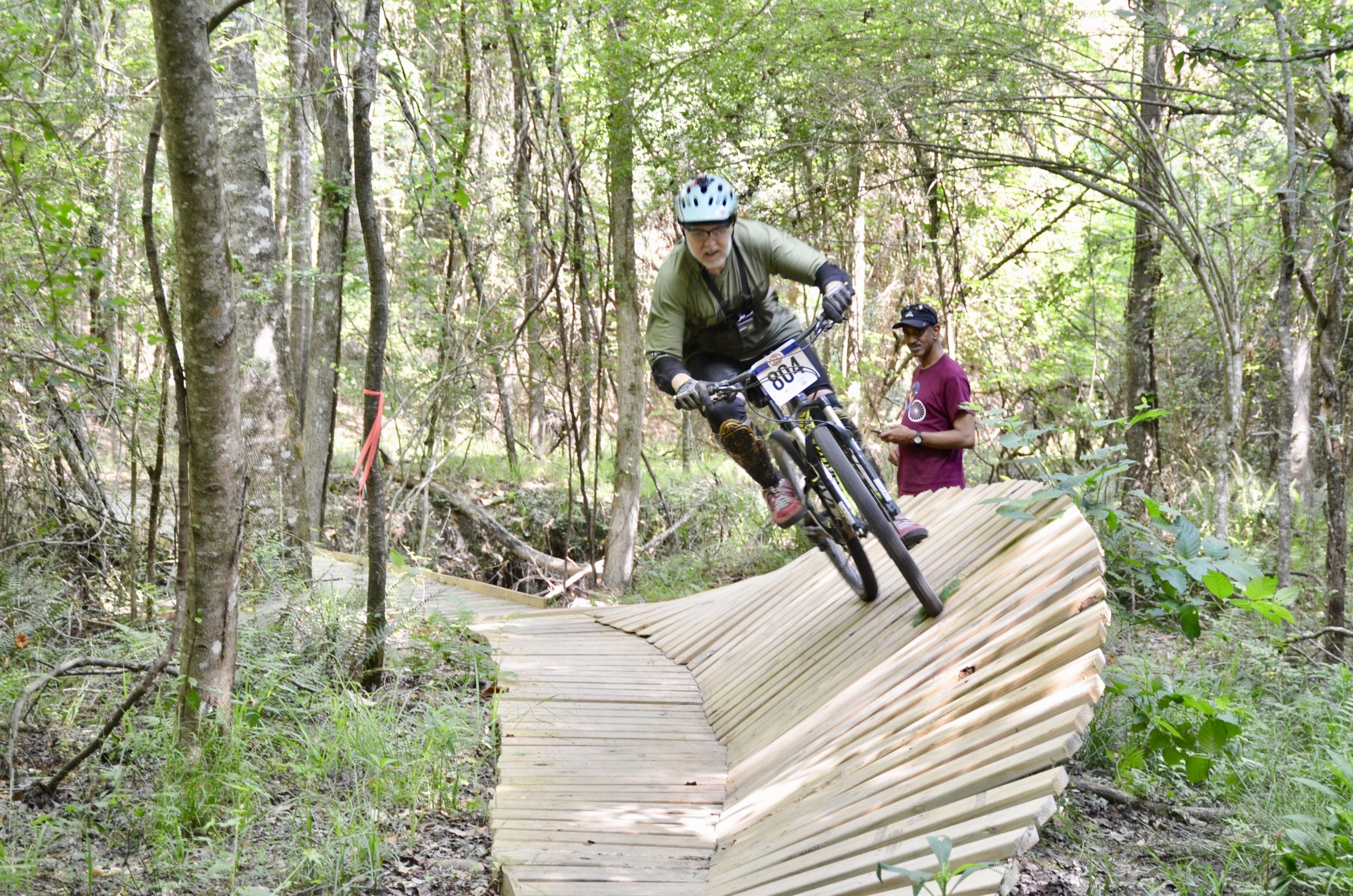 A mountain biker navigates a wooden ramp in a forested area. The rider, wearing a helmet and sporting number 804 on their jersey, is captured mid-jump, showcasing their skills. In the background, another spectator watches the action. The scene is surrounded by trees and greenery, highlighting the natural environment of the biking trail. Mt. Zion Bike Trails mountain bike trail.