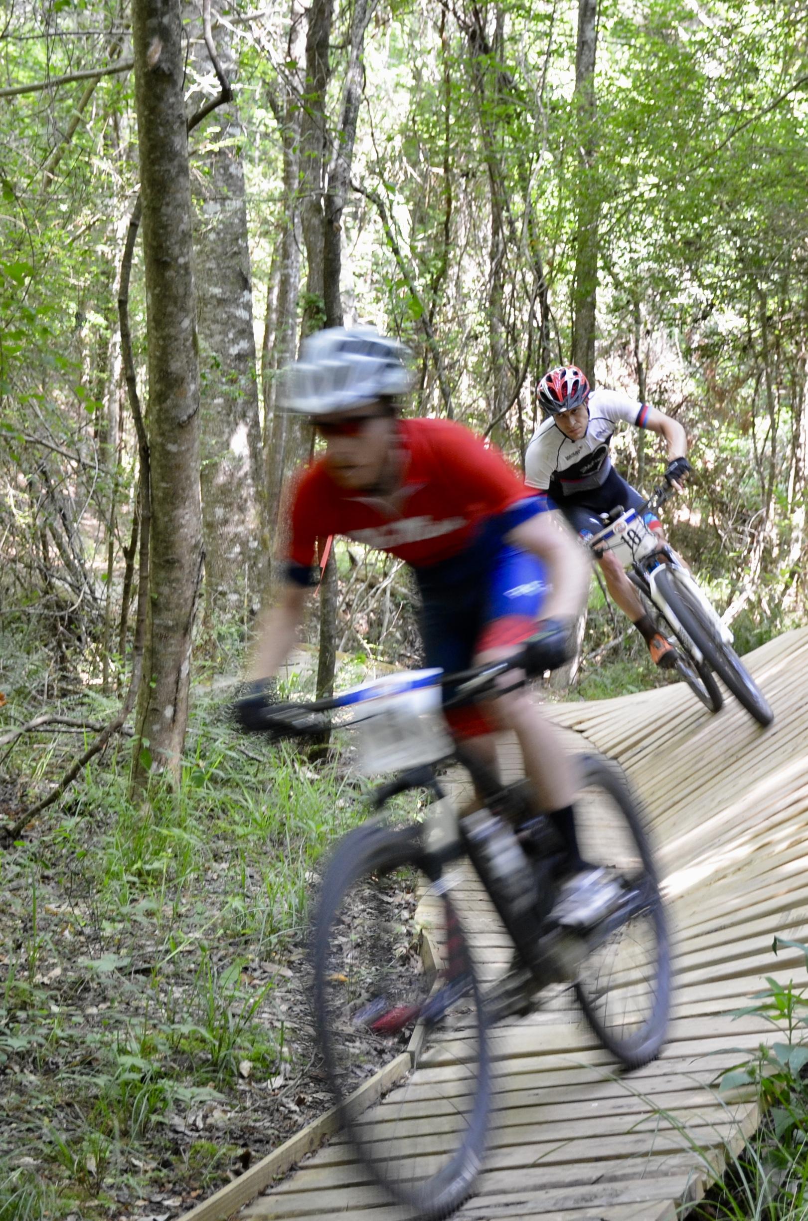Two mountain bikers racing on a wooden ramp in a forested area, with one rider in a red shirt and the other in a white shirt. The image captures the action with a slight motion blur, emphasizing their speed as they navigate the trail surrounded by trees and greenery. Mt. Zion Bike Trails mountain bike trail.