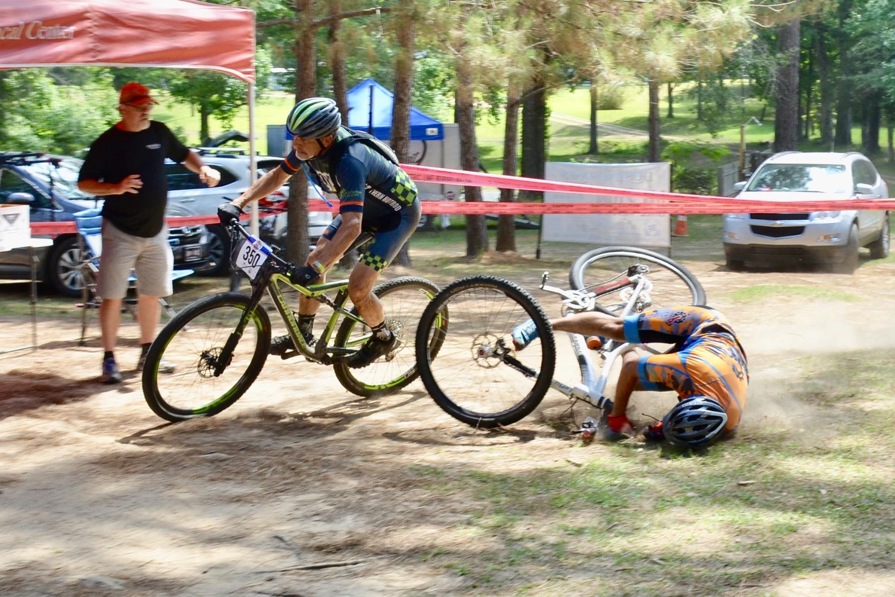 A mountain biker in a blue and green jersey navigates a turn, while another cyclist in an orange jersey has fallen off their bike onto the ground, raising a cloud of dust. In the background, a spectator in a black shirt appears concerned, and several vehicles and tents are visible, indicating a biking event. Mt. Zion Bike Trails mountain bike trail.
