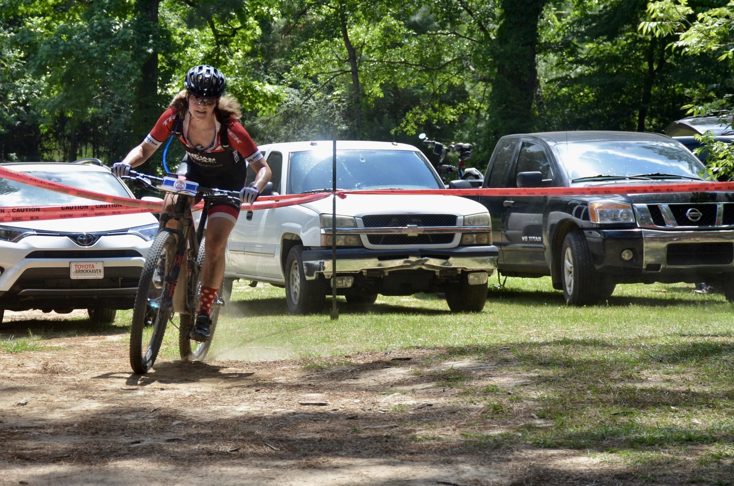 A mountain biker wearing a black helmet and a red and black cycling outfit races along a dirt path, navigating between parked vehicles and caution tape. Dust is kicked up behind her as she approaches the end of a course in a wooded area. Mt. Zion Bike Trails mountain bike trail.