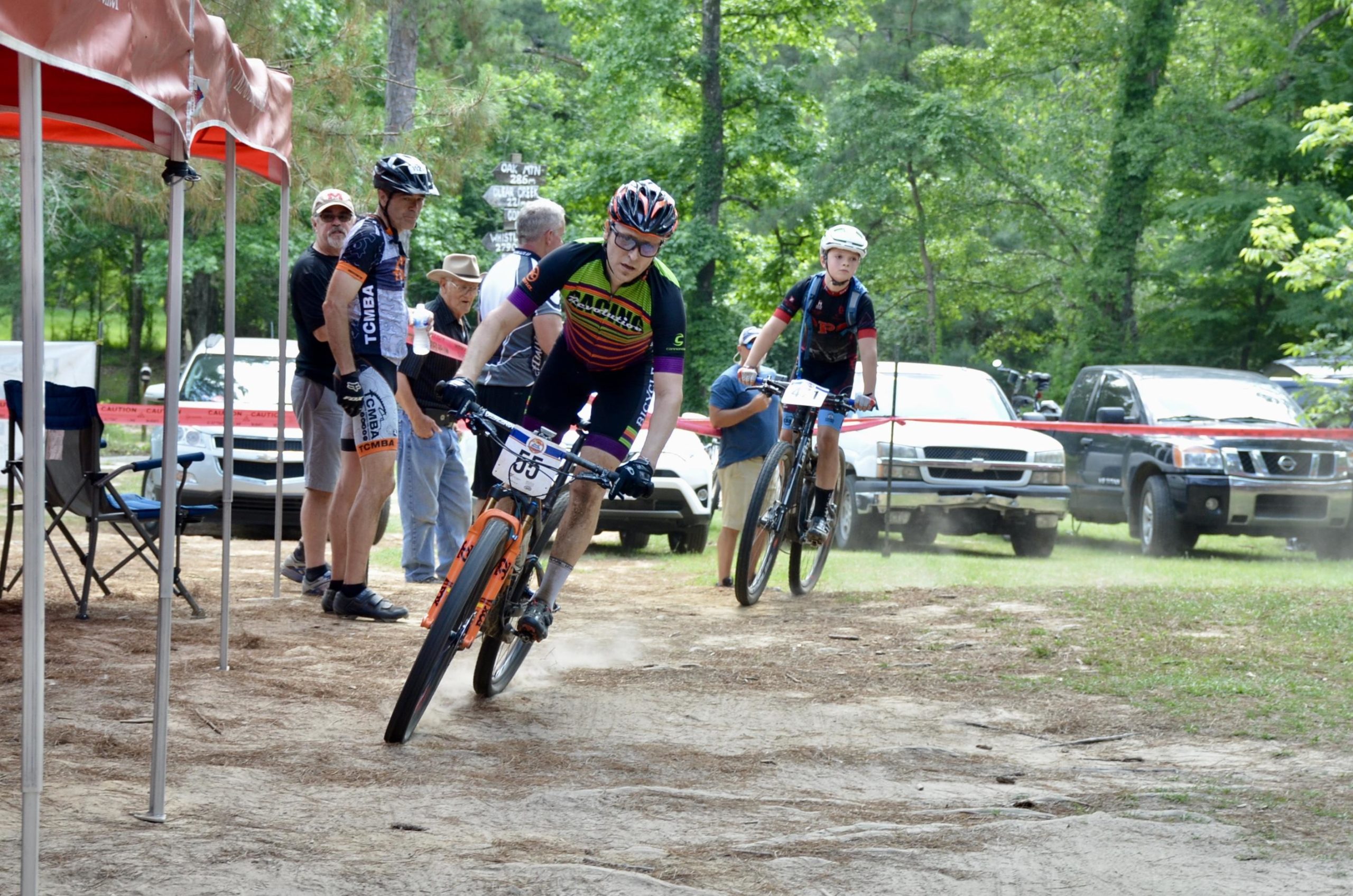 A group of mountain bikers competes in an outdoor cycling event. In the foreground, an adult male cyclist is maneuvering a bike on a dirt path, while a young cyclist follows closely behind. Spectators are gathered nearby, some under a red tent, observing the race. Trees and parked vehicles can be seen in the background, creating a lively, competitive atmosphere. Mt. Zion Bike Trails mountain bike trail.