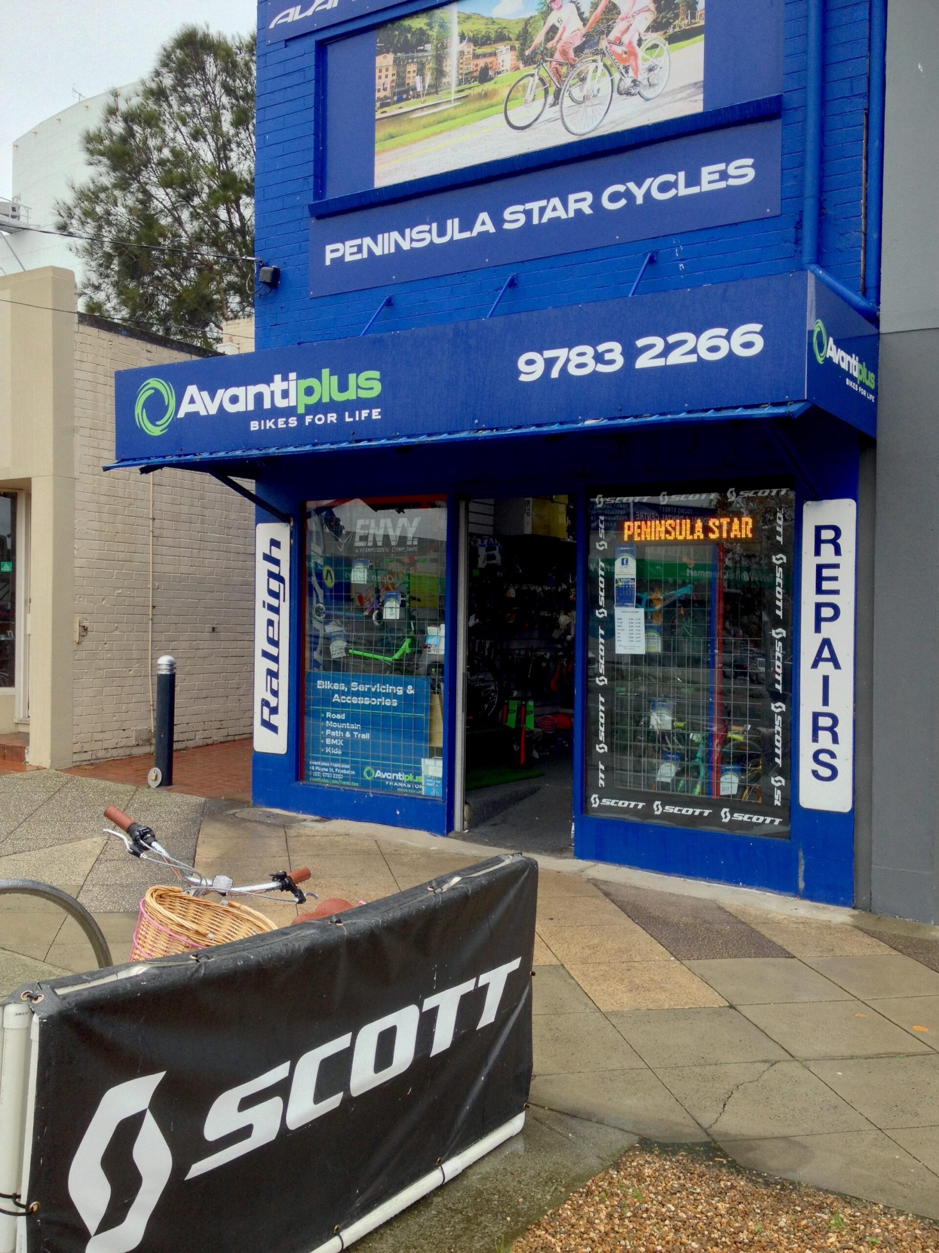A storefront of a bicycle shop named "Peninsula Star Cycles," featuring bright blue walls and signage. The shop displays a range of bikes and accessories. A bicycle with a basket is positioned in front of a black barrier advertising "Scott" products. The atmosphere is inviting, with a focus on cycling services and sales.