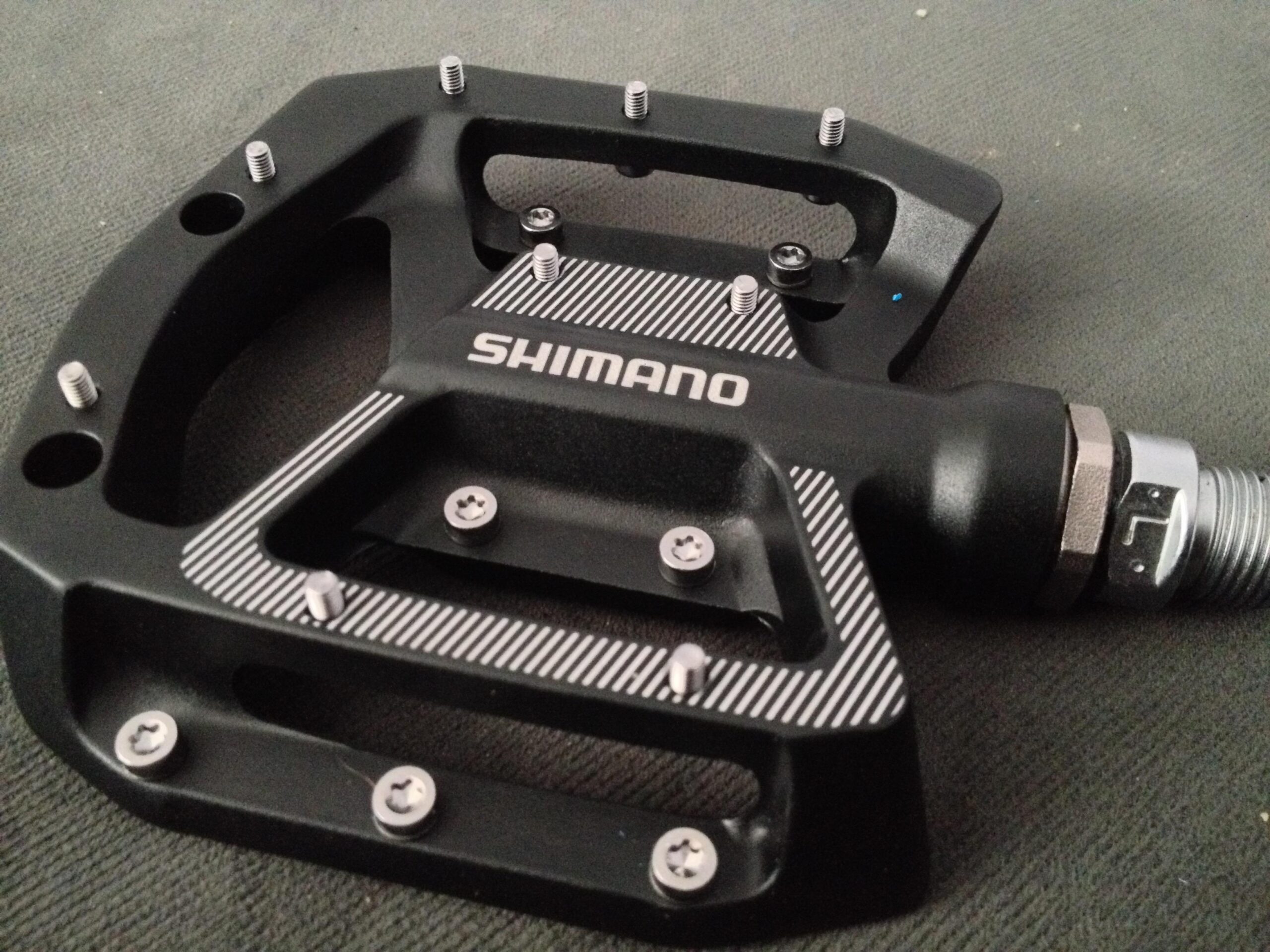 A close-up view of a Shimano bicycle pedal, featuring a black metal body with silver screws and a textured grip surface. The pedal has a flat design with an integrated connector for attachment to a bike.
