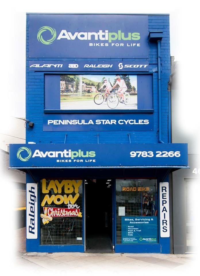 Alt text: Exterior of a bike shop named "Avantiplus Bikes for Life," featuring a blue facade, promotional signage for brands like Avanti, Raleigh, and Scott, and a large display screen showcasing cyclists. The entrance has a colorful sign that reads "Layby Now for Christmas." The shop offers bike repairs and accessories.