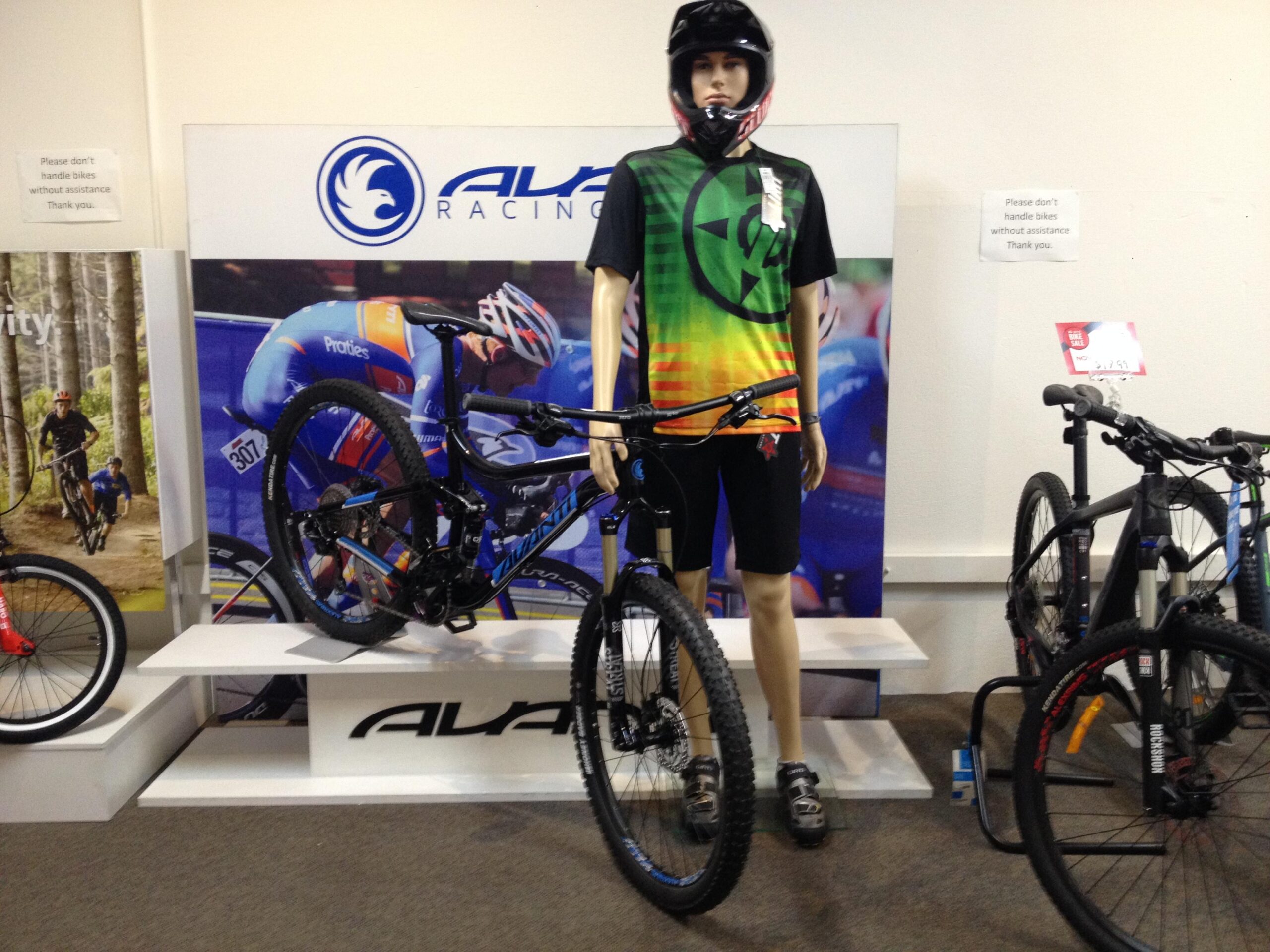 A mannequin dressed in a colorful cycling jersey and helmet stands beside a mountain bike on display. The backdrop features a racing scene, and a sign advises customers not to handle bikes without assistance. Additional bicycles are visible in the foreground.