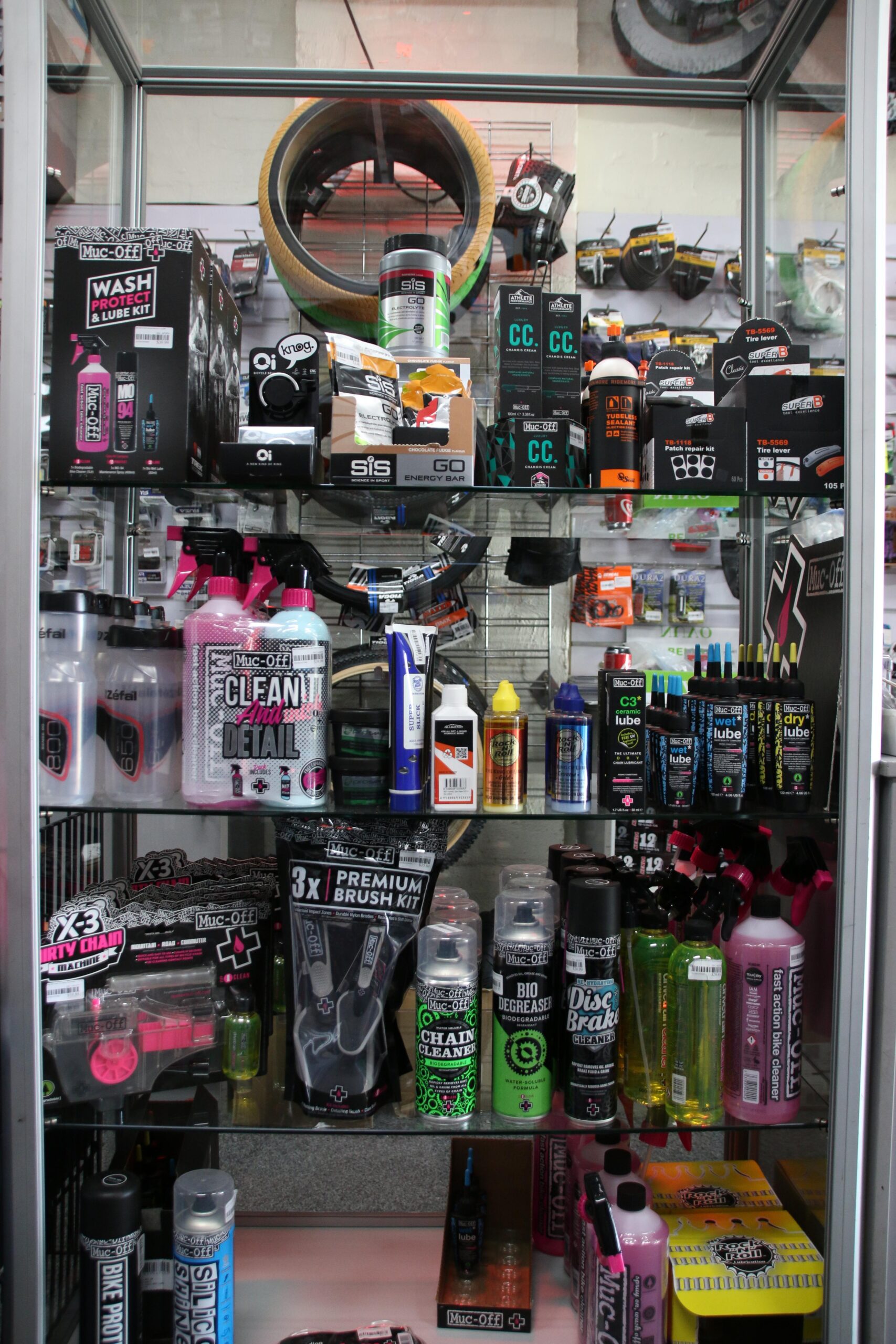 A glass display case filled with various bicycle maintenance products and cleaning supplies. The items include cleaning sprays, lubricants, detailing kits, energy bars, brush kits, and tire tools. Prominent brands like Muc-Off and SIS are featured prominently on the packaging. In the background, colorful bike tires and accessories are visible.