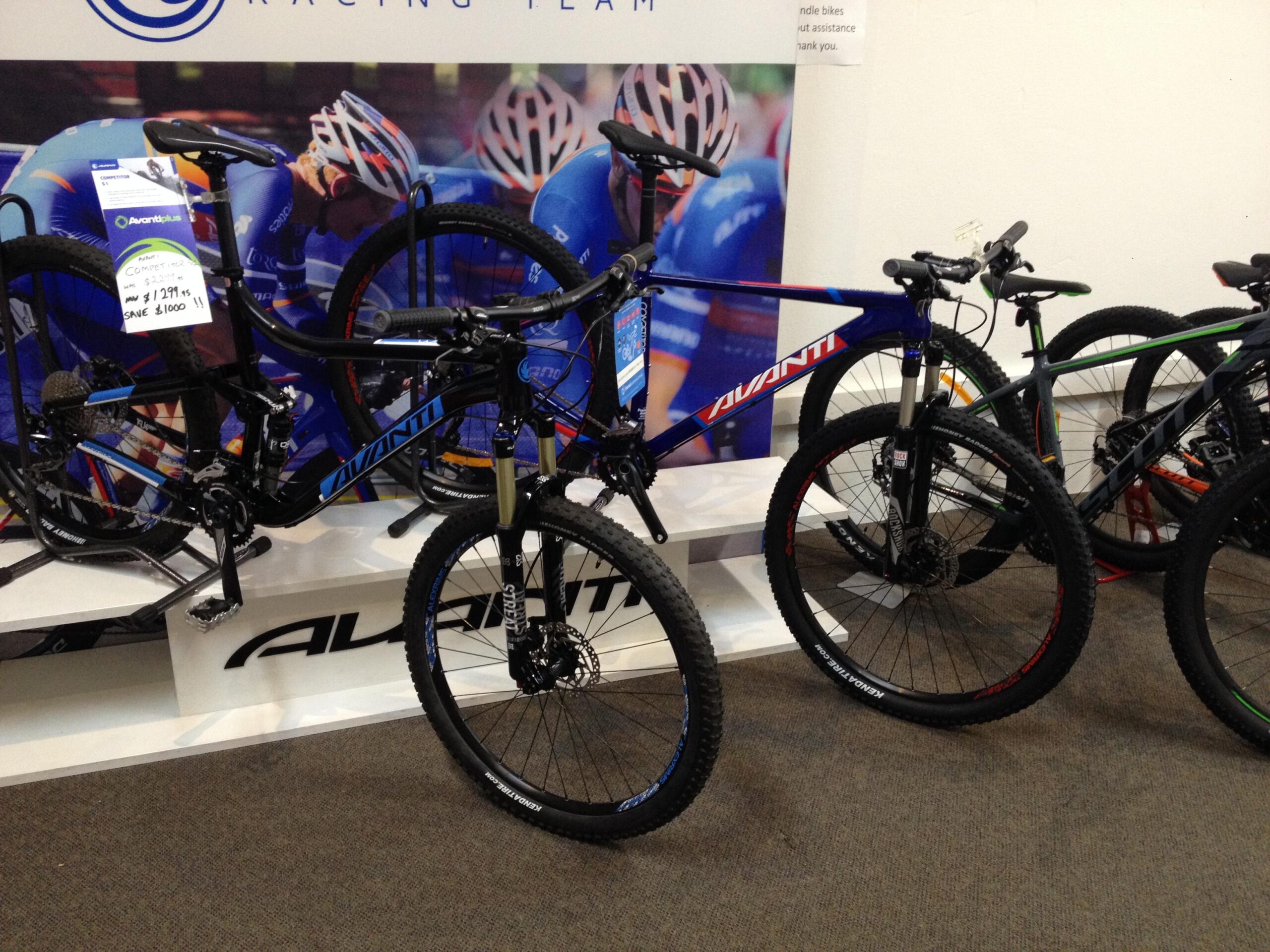 Two mountain bikes are displayed on a shelf in a shop setting. One bike is predominantly black with blue accents, while the other features a blue and red color scheme. Both bikes have thick tires, designed for rugged terrain. Price tags are visible, indicating discounts. In the background, a poster of cyclists in action is partially visible, along with the Avanti logo.