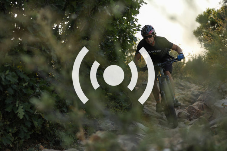 A mountain biker navigates a rocky trail surrounded by greenery, wearing a helmet and gloves, and focused on the path ahead. An icon representing sound or live broadcasting is overlaid in the center of the image.