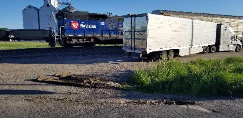 A blue freight train with the logo "Rail Link" parked next to a white semi-truck trailer on a gravel road, surrounded by green grass and industrial buildings in the background. The train and truck are positioned near a track, with some debris visible on the road.
