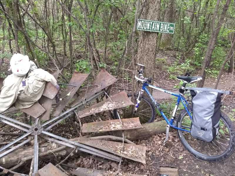 A mountain bike parked beside an old rusted piece of machinery, partially covered by greenery. A sign reading "Mountain Bike Trail" is visible on a tree in the background. A figure dressed in beige clothing is seated on the equipment, with a white mask visible. Elm Creek Park mountain bike trail.