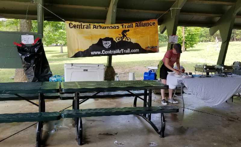 A person preparing food at a picnic shelter, with a banner reading "Central Arkansas Trail Alliance" in the background. Several picnic tables are visible, and there are various items and equipment set up for an outdoor event. The setting is green and wooded, suggesting a park environment. Boyle park mountain bike trail.