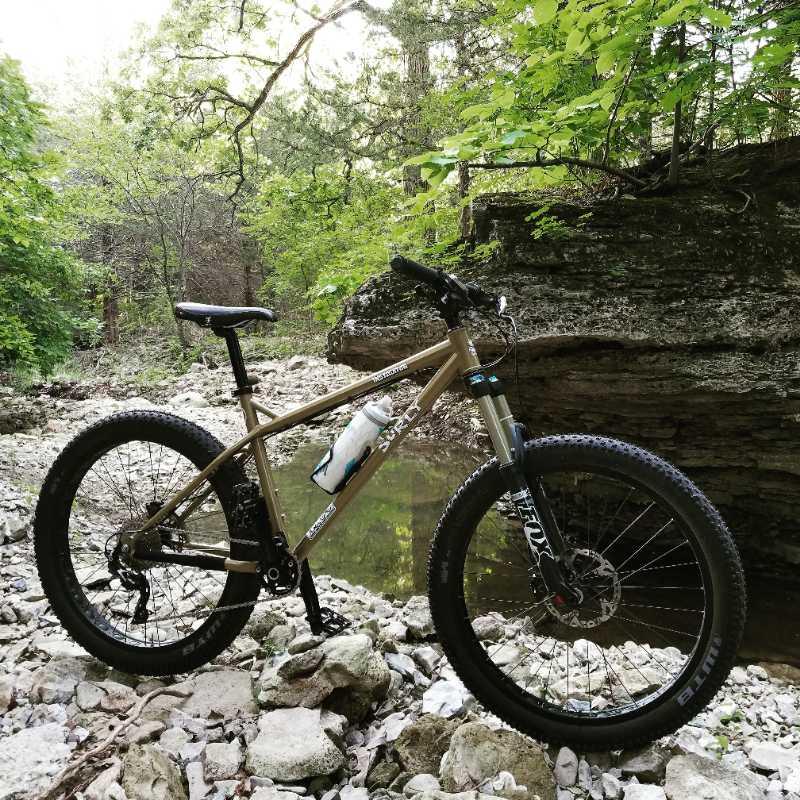 A mountain bike rests on rocky terrain beside a small body of water, surrounded by lush green foliage and trees. The bike is equipped with a water bottle mounted on the frame, and the landscape features a mix of stones and grass, creating a natural outdoor setting. Alcove Springs mountain bike trail.