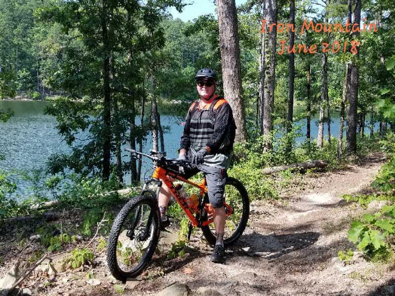 A person wearing a helmet and cycling gear stands beside an orange mountain bike on a trail near a lake. The background features trees and a scenic landscape, indicating a sunny day in Iron Mountain. The text overlay indicates that the photo was taken in June 2018. Iron Mountain mountain bike trail.