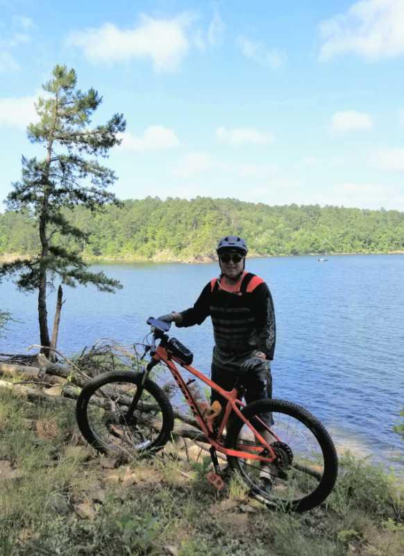 A person in biking attire stands next to an orange mountain bike near a peaceful lake, surrounded by trees and lush greenery under a partly cloudy sky. Iron Mountain mountain bike trail.