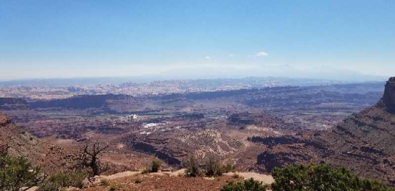 A panoramic view of a vast desert landscape under a clear blue sky. The foreground features rocky terrain with sparse vegetation, while rolling hills and valleys extend into the distance, showcasing a mix of earthy tones. Farther back, a faint outline of mountains is visible. The scene captures the beauty of nature and the expansive view from a high vantage point. Big Chief mountain bike trail.
