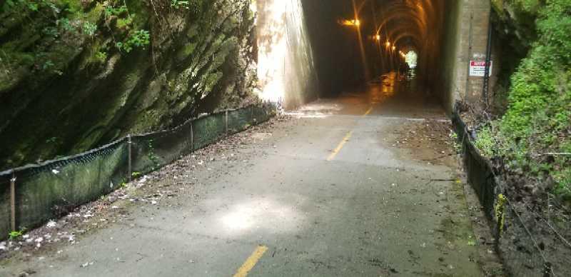 A narrow, dimly lit tunnel with rocky walls and a paved path running through it. The pathway is lined with fencing and leads into darkness, illuminated by sparse overhead lights. Green foliage is visible on the tunnel's entrance and sides, suggesting a natural setting. A caution sign is posted near the entrance, indicating potential safety concerns. Silver Comet Rail Trail mountain bike trail.