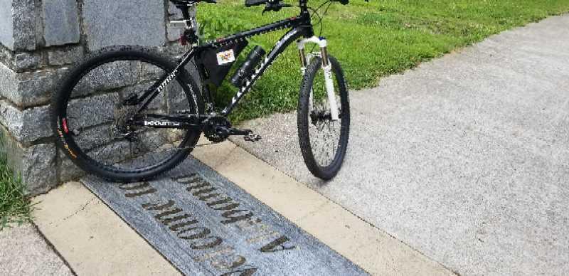 A black mountain bike leaned against a stone pillar next to a paved walkway, with a sign engraved on a grey stone slab that reads "We Come To 4-Joy, 4-Humanity." Lush green grass is visible in the background. Silver Comet Rail Trail mountain bike trail.